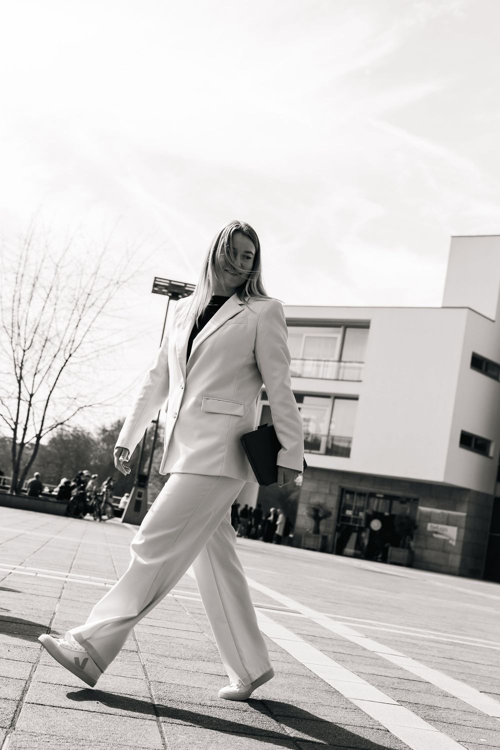 Black and white photo of a blonde woman in a white pantsuit walking confidently across a city street.