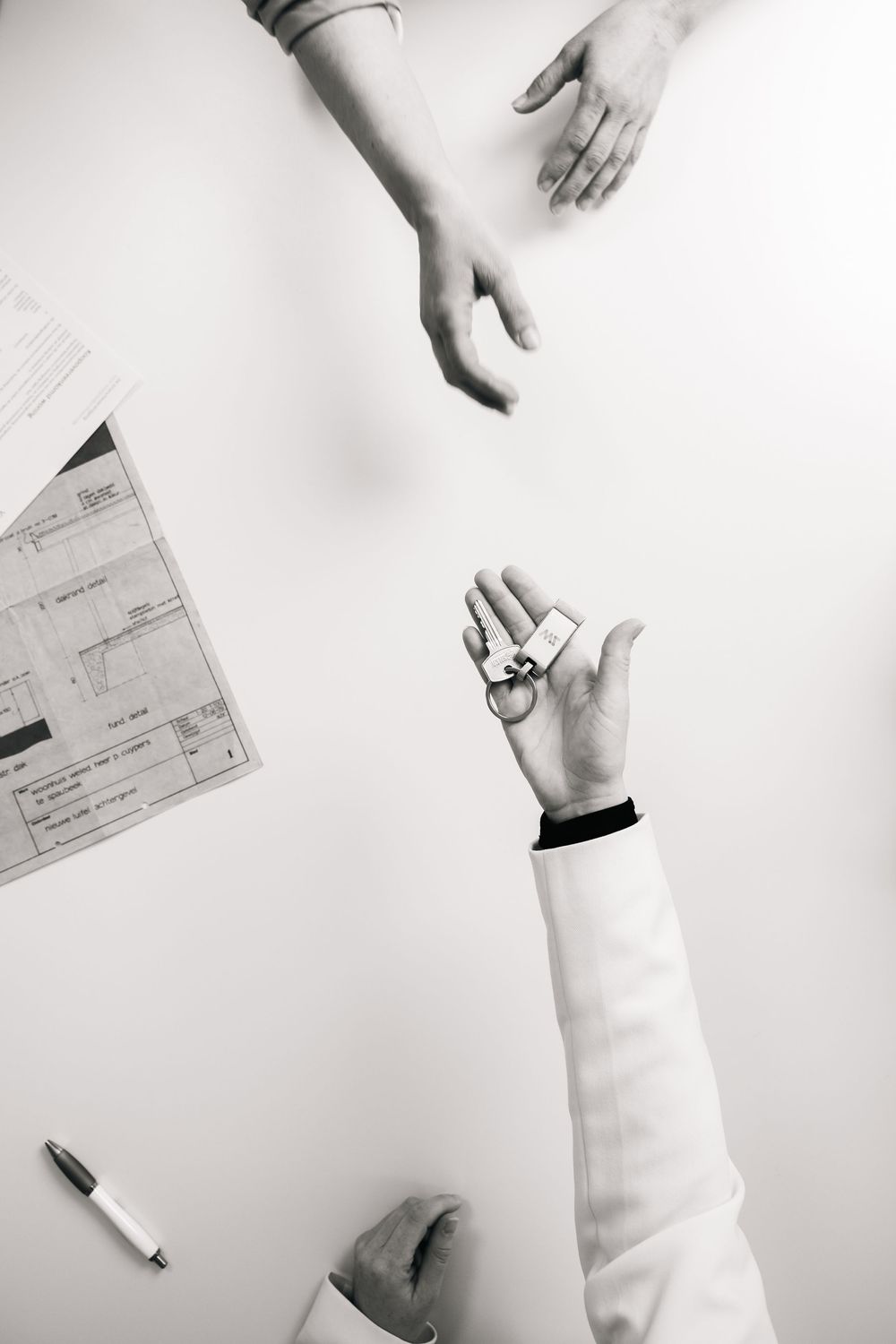 Overhead black and white photo of hands exchanging keys over a desk with documents and a pen.