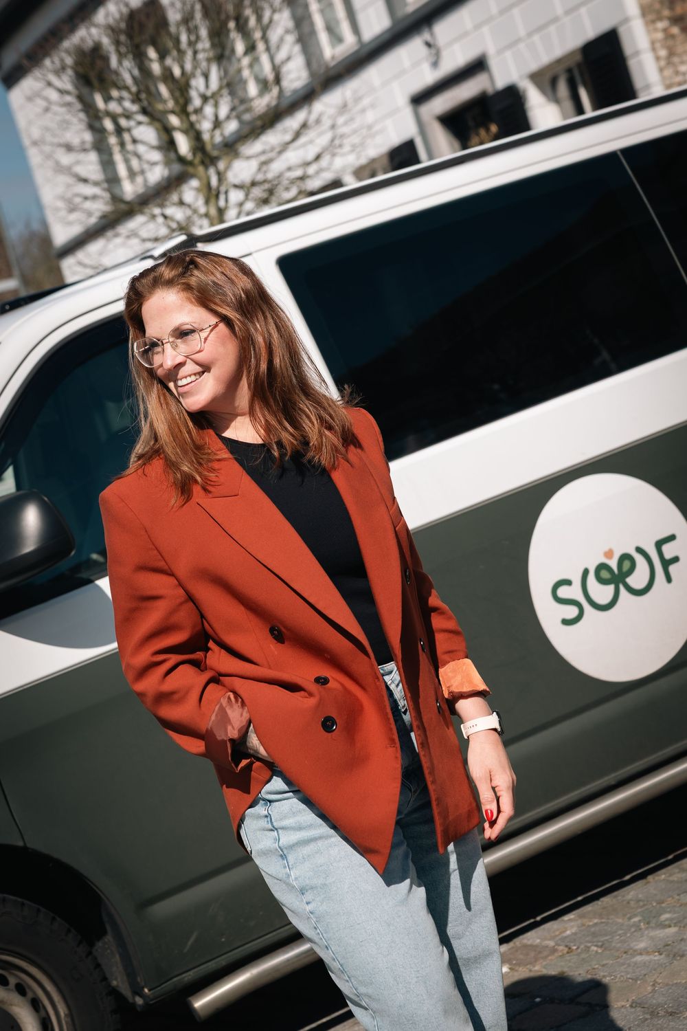 Woman in red blazer and jeans smiling outdoors in front of a SOOF branded van on a sunny day.