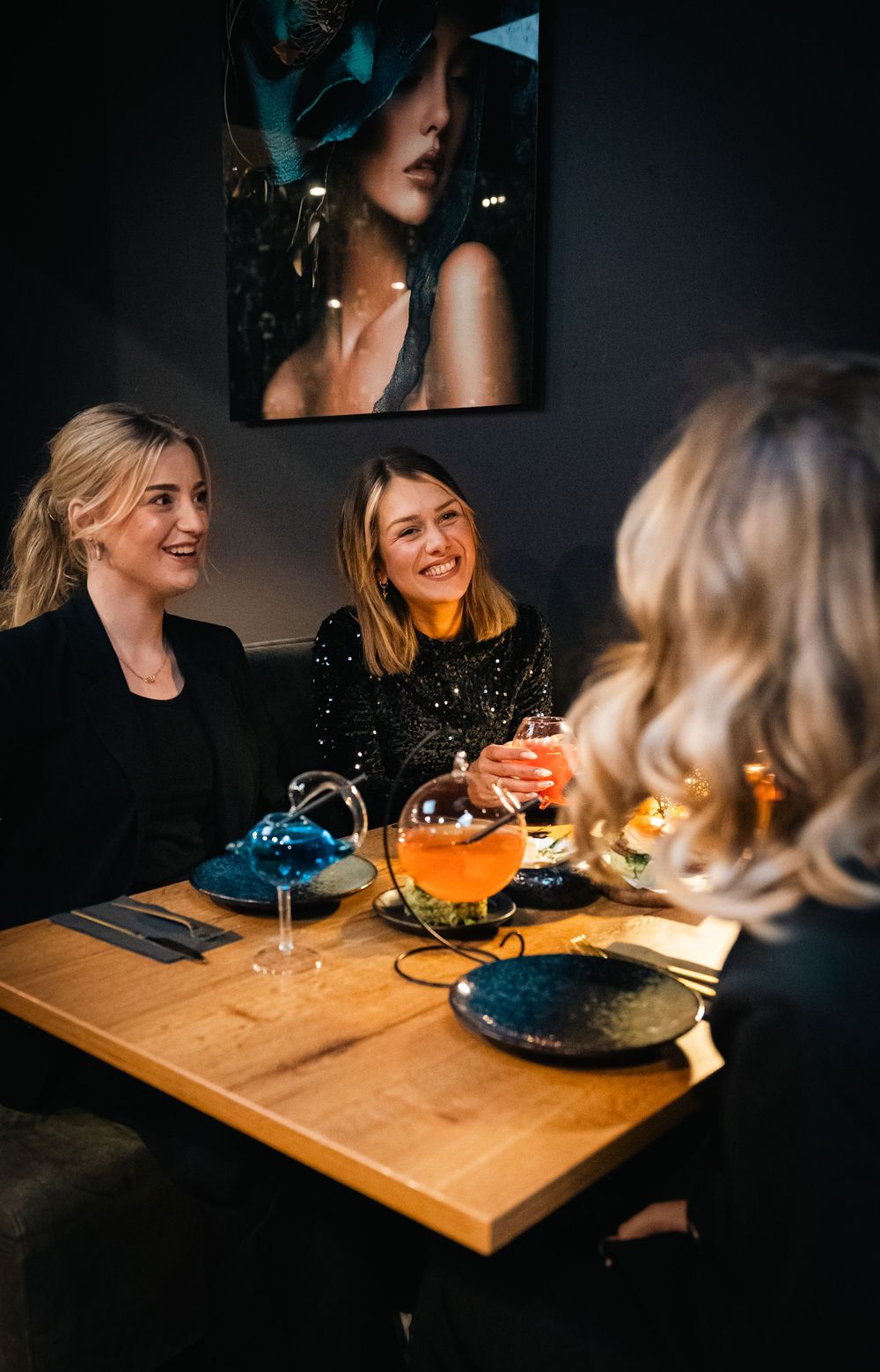 Three women in black outfits laughing over cocktails at a dimly lit restaurant table with elegant decor.
