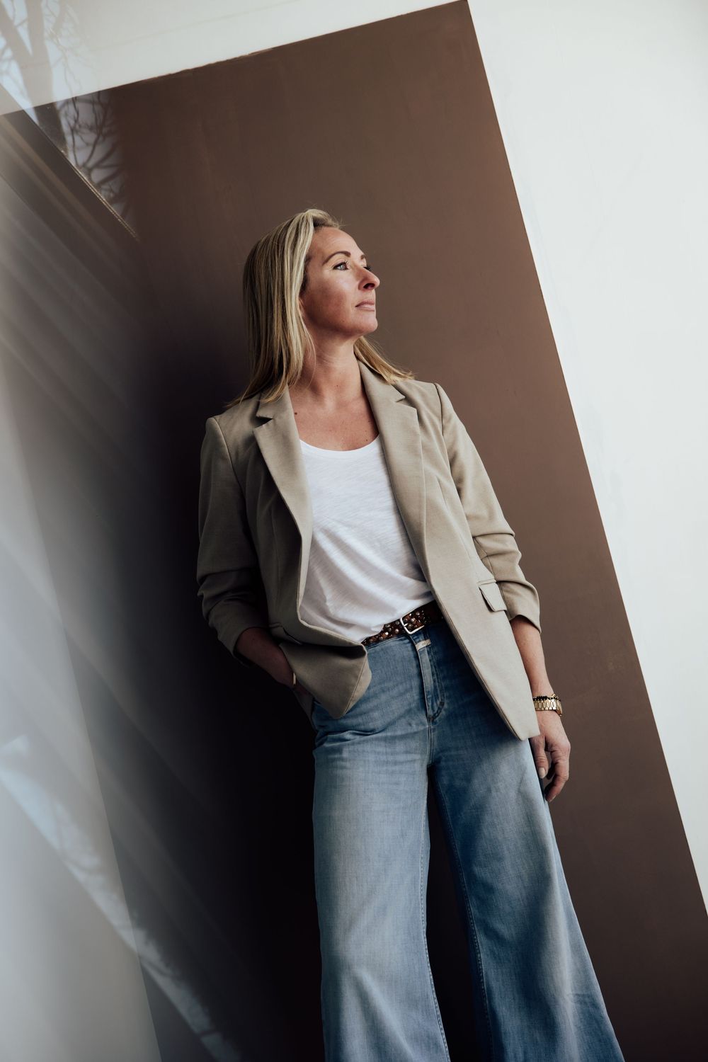 Blonde woman in beige blazer, white tee, and wide-leg jeans leaning against a brown wall, looking upward.