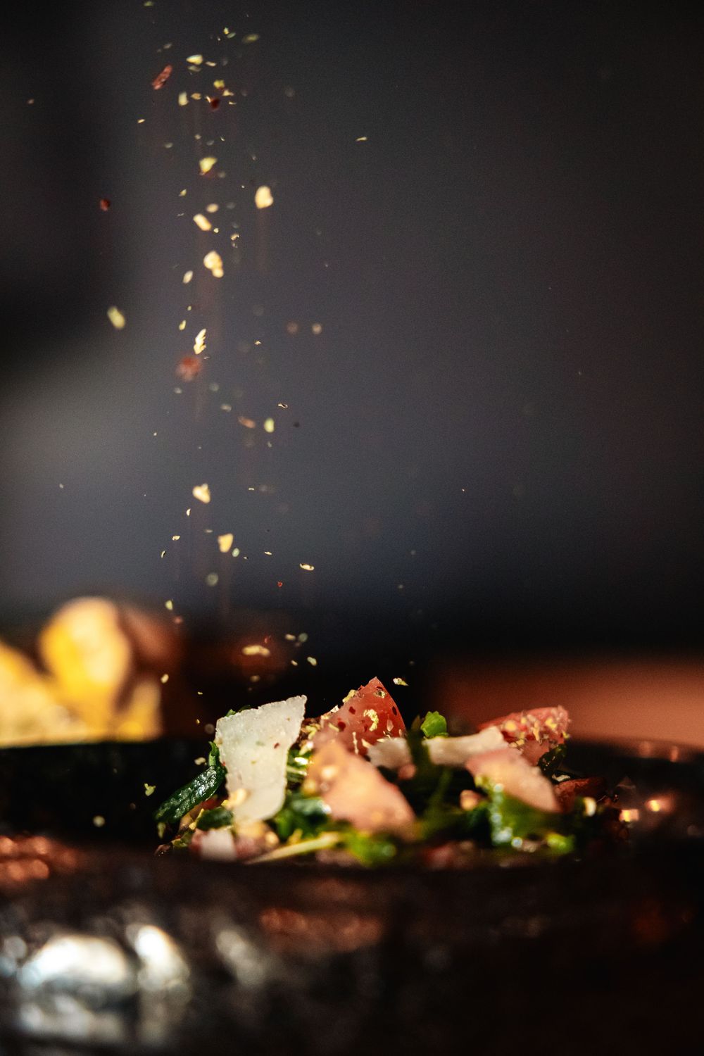 Spices falling onto a gourmet salad with greens, tomatoes, and parmesan on a dark surface.