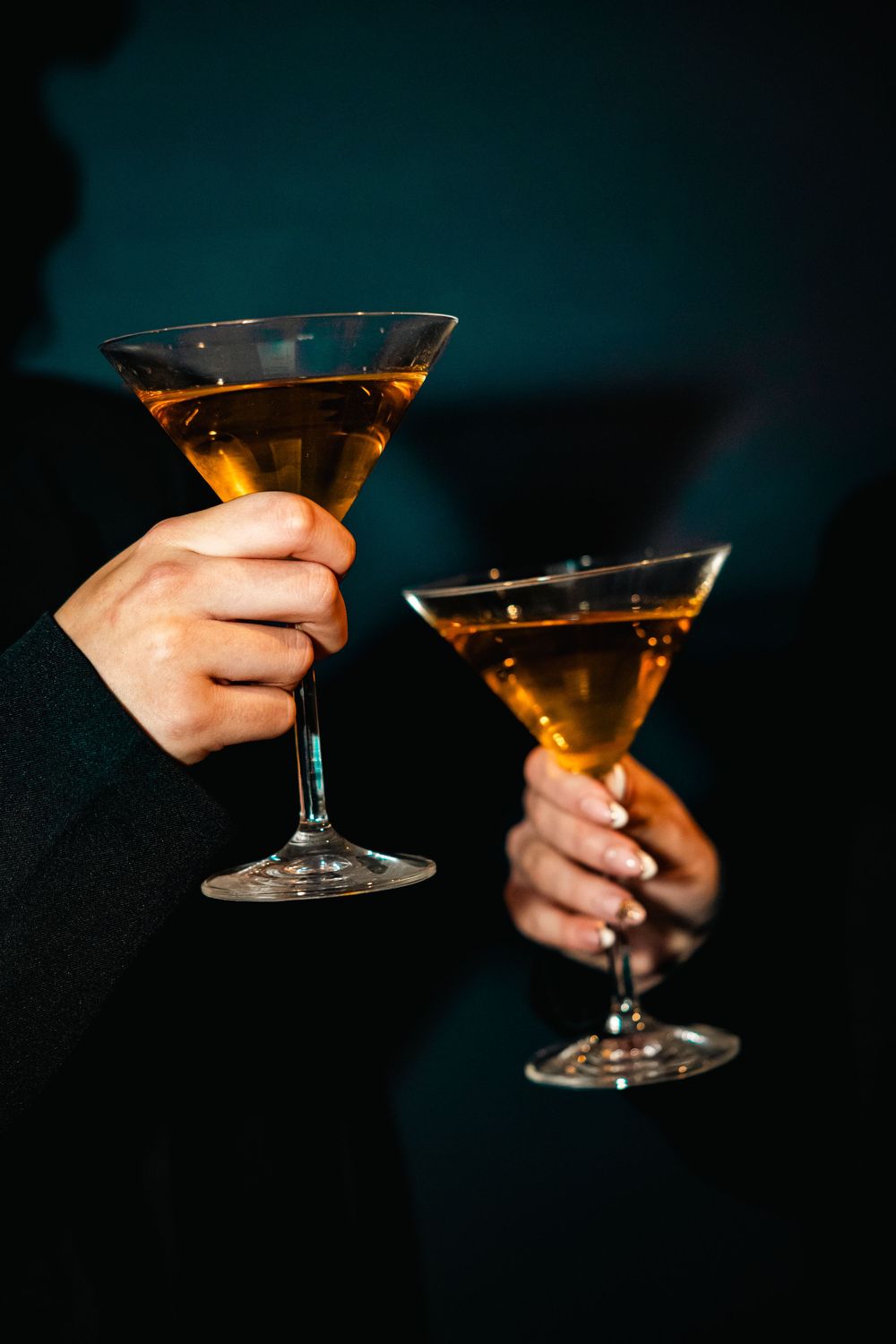Two hands holding amber cocktails in martini glasses at an elegant evening event.