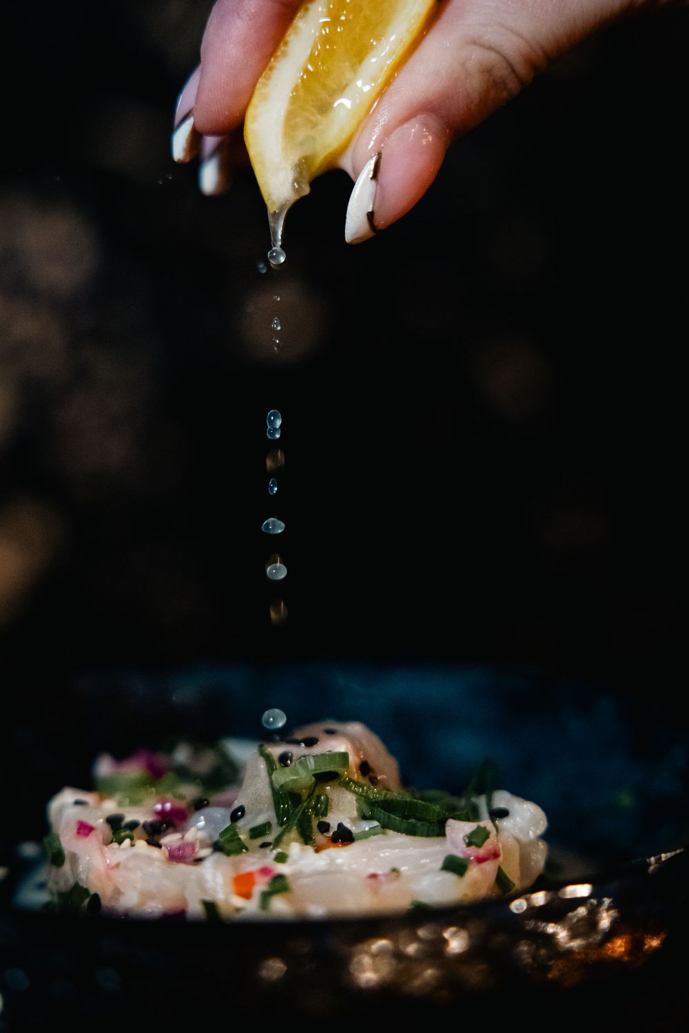 Hand squeezing fresh lemon juice over seasoned fish fillet on a dark cooking surface.