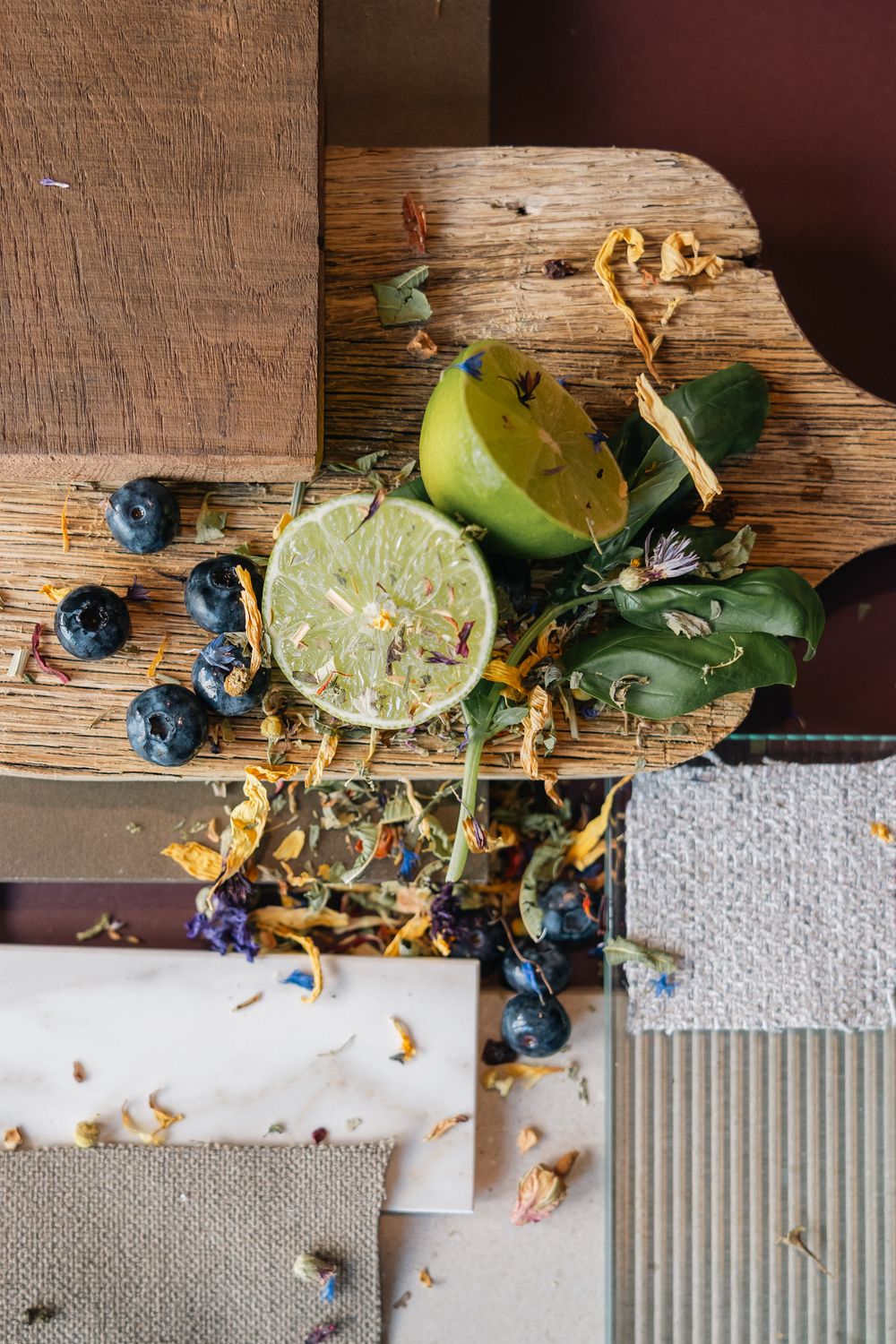 A rustic wooden cutting board with fresh lime, blueberries, dried flowers, and tea leaves arranged on layered material swatches.