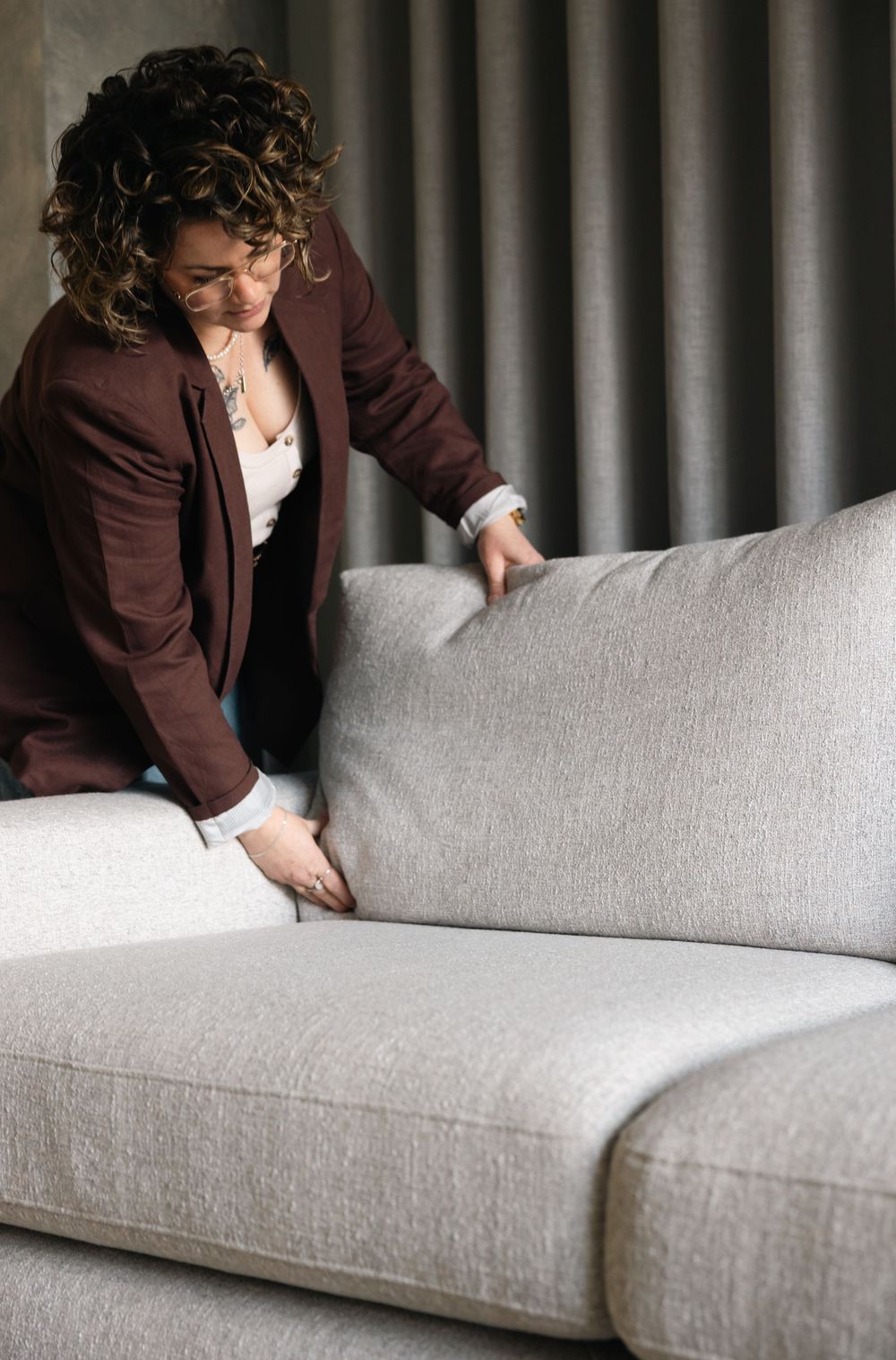 Woman in burgundy blazer arranging cushions on a modern light gray fabric sofa indoors.
