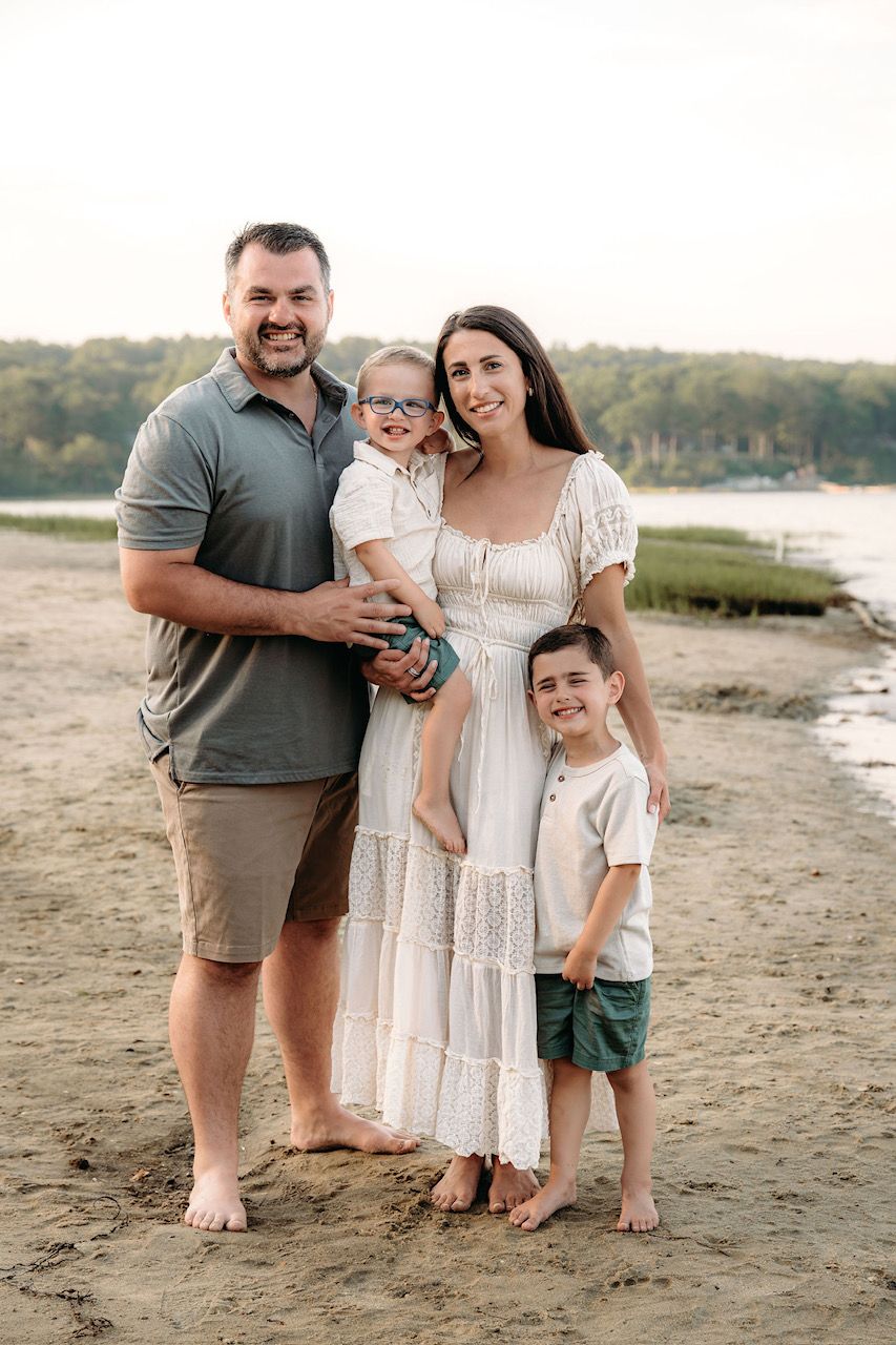 Family of four posing on sandy beach at sunset, parents holding young children, wearing casual summer outfits.