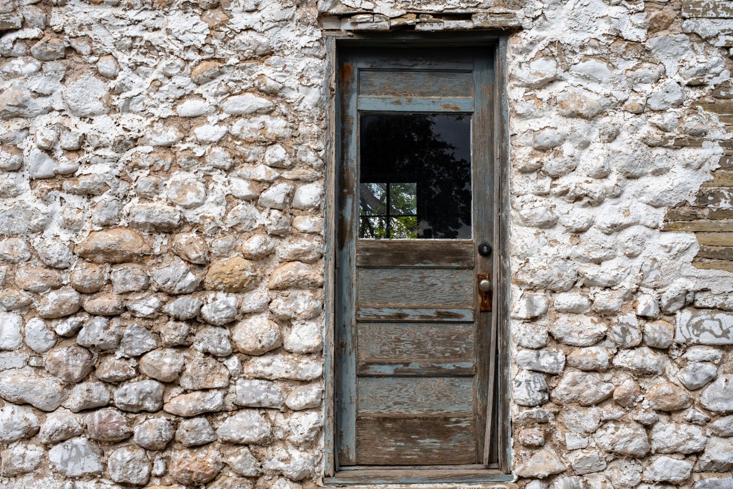 Weathered wooden door with glass panel set in rustic limestone stone wall of an old abandoned building.