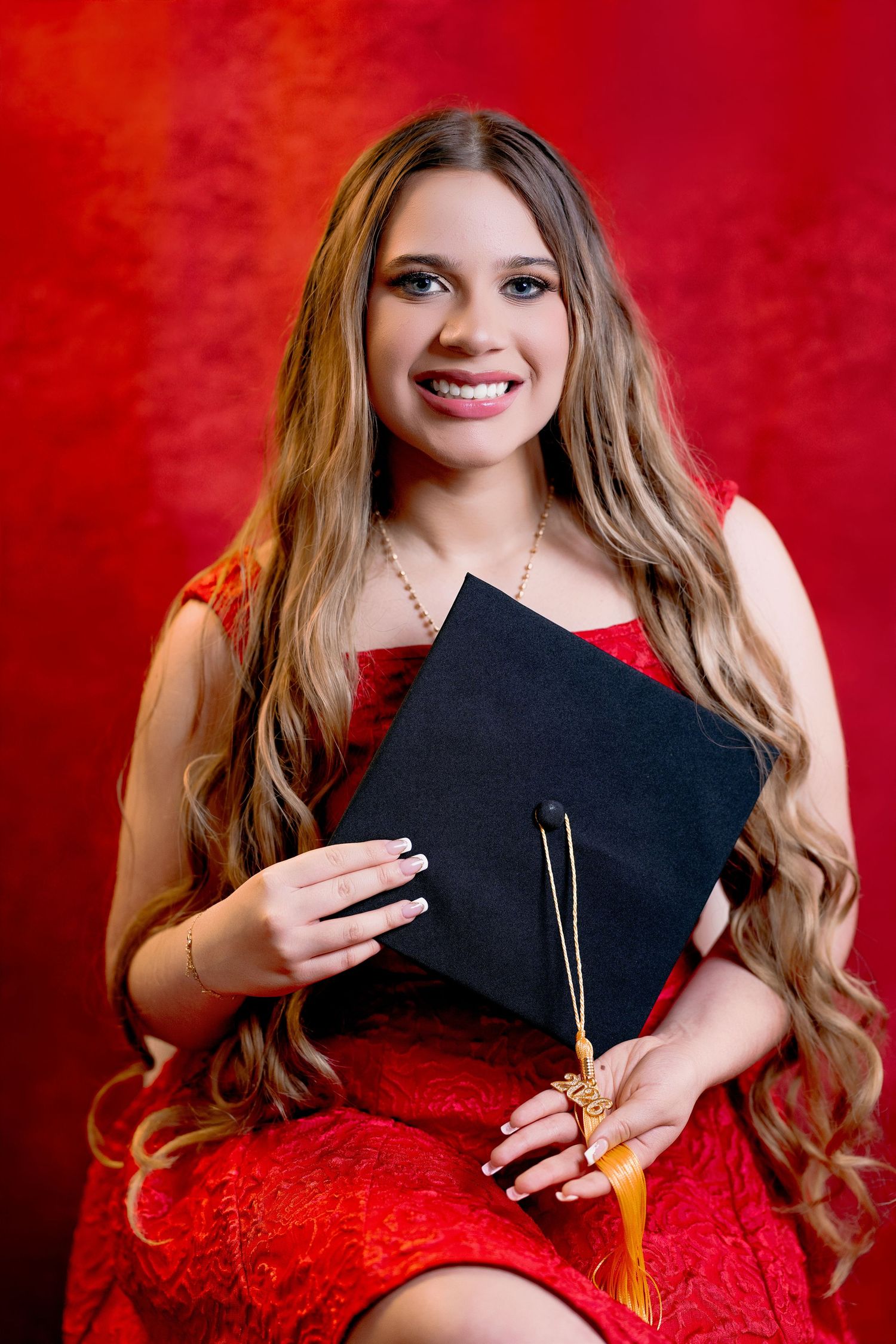 Smiling young woman in red dress holding black graduation cap with gold tassel against red background.