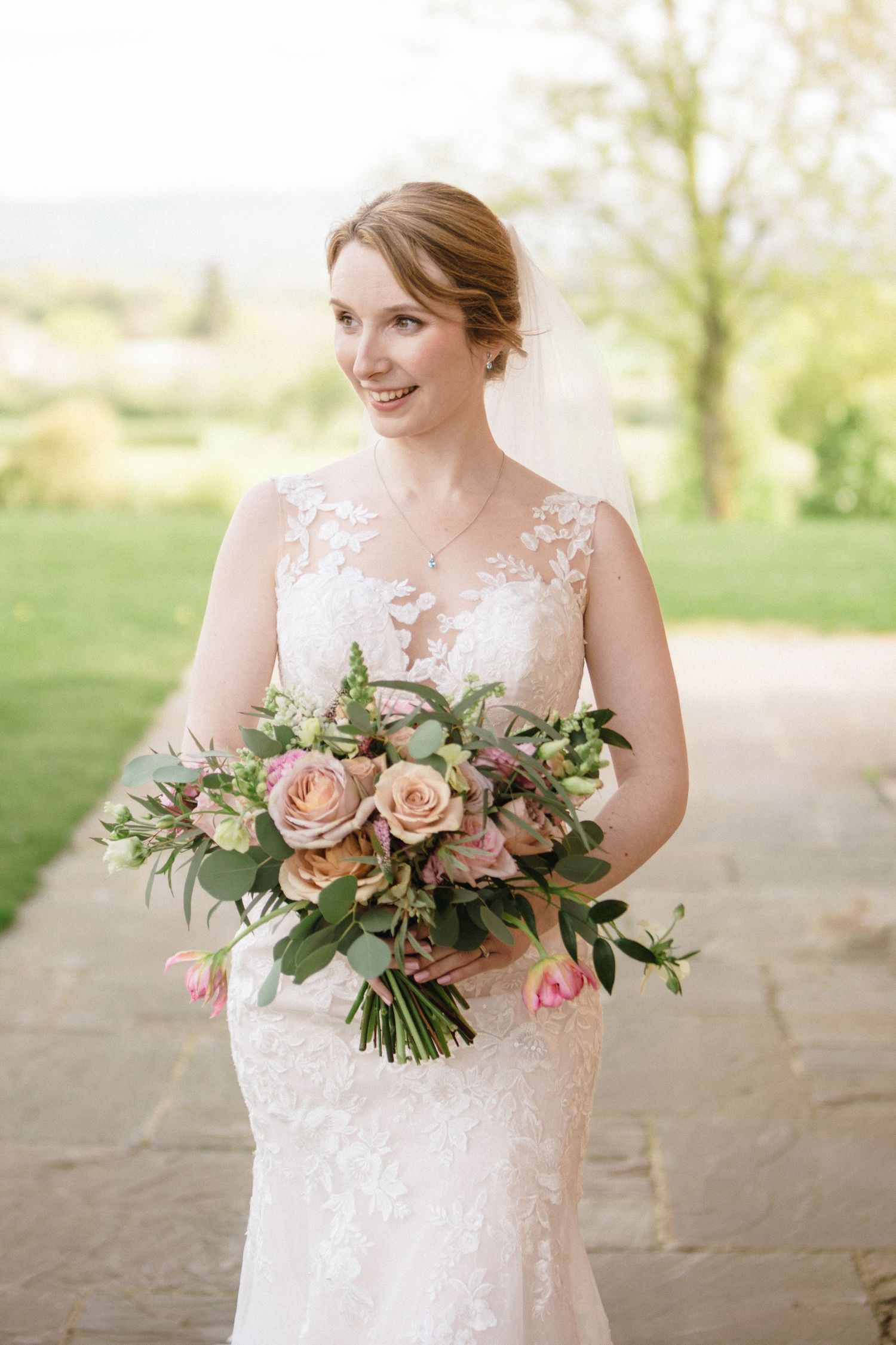 Smiling bride in lace wedding dress holding a pink and peach rose bouquet with greenery outdoors.