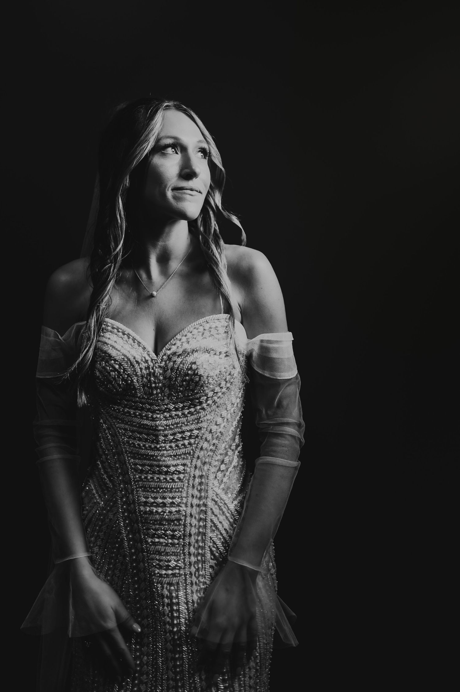 Elegant bride in beaded off-shoulder gown poses dramatically in black and white portrait.