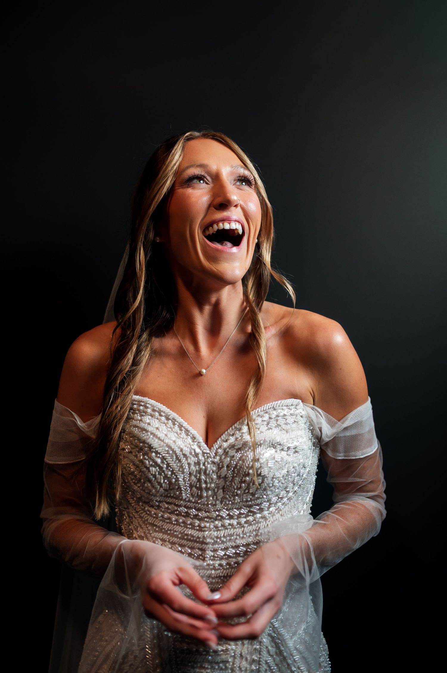 A joyful bride in a beaded off-shoulder gown laughs radiantly against a dark studio background.