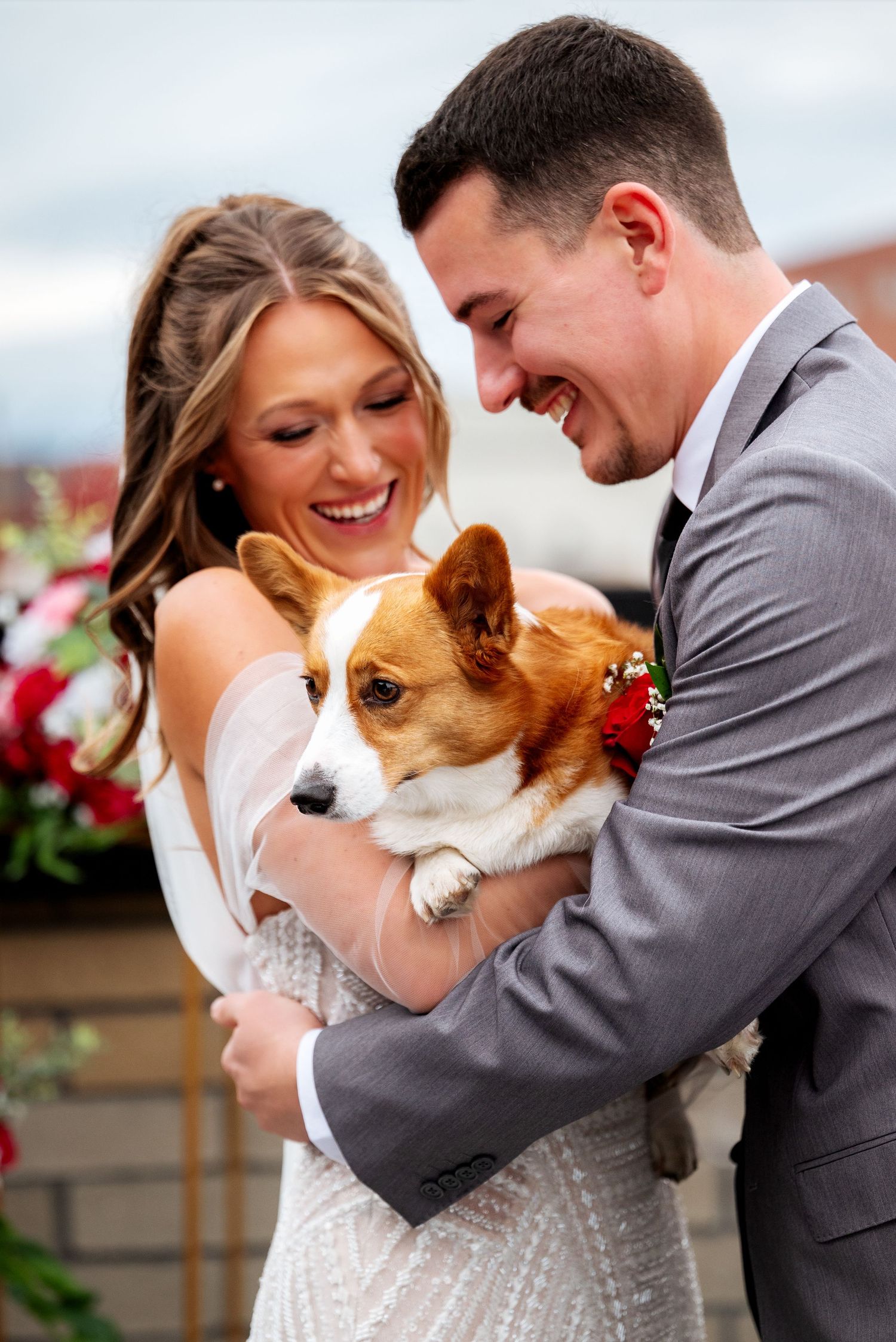 Smiling bride and groom holding an adorable Corgi dog on their wedding day.