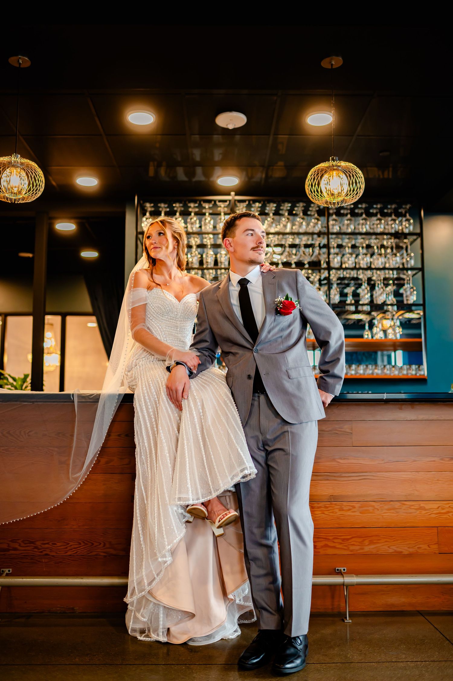 Bride in white gown and groom in gray suit pose together at a bar during their wedding reception.