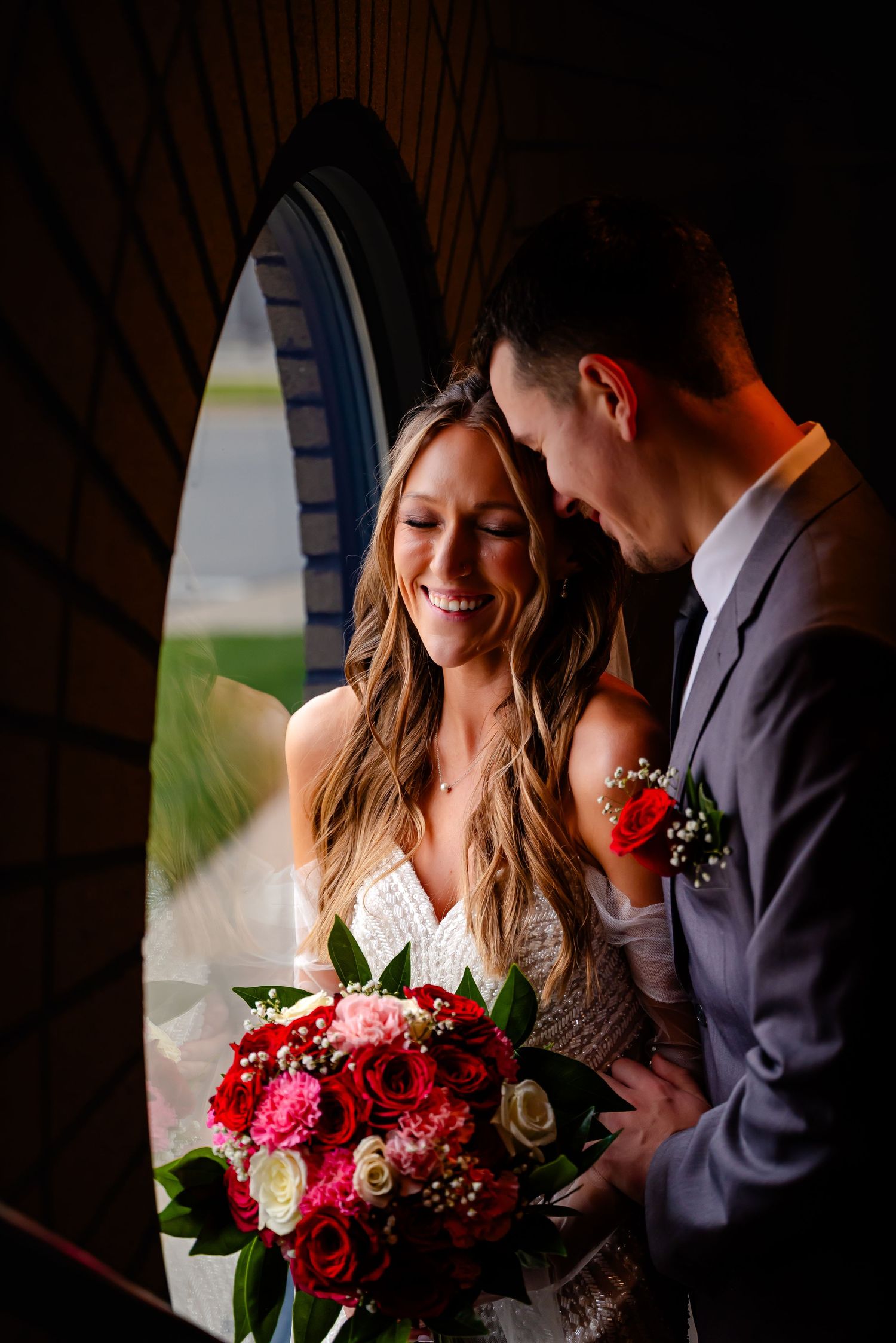 Smiling bride holding pink and red rose bouquet with groom in gray suit sharing romantic moment in arched doorway.