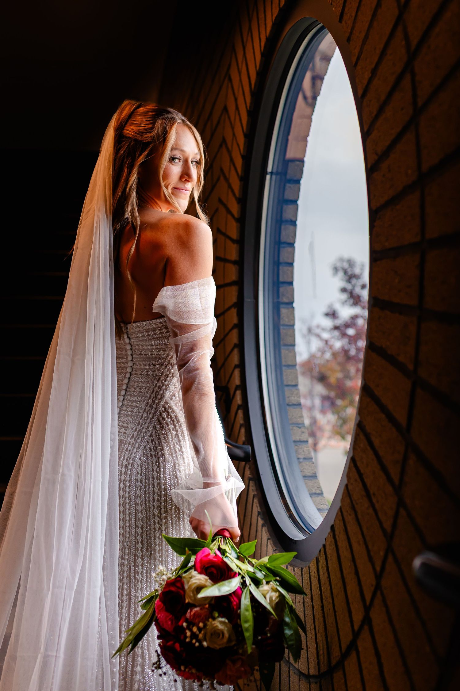 Bride in strapless wedding gown and veil holds bouquet by round window.