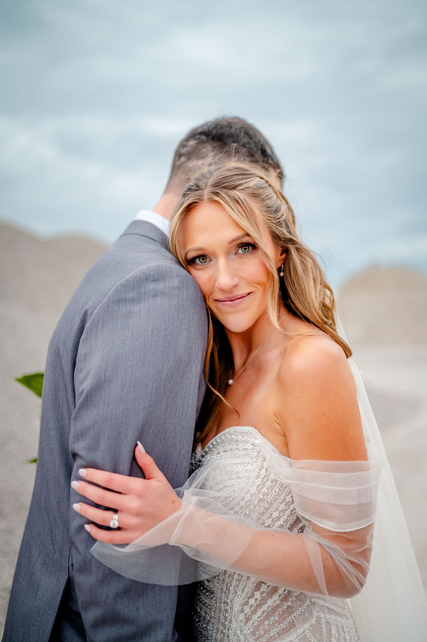Bride in beaded off-shoulder gown and veil embraces groom in gray suit on sandy beach, smiling at camera.