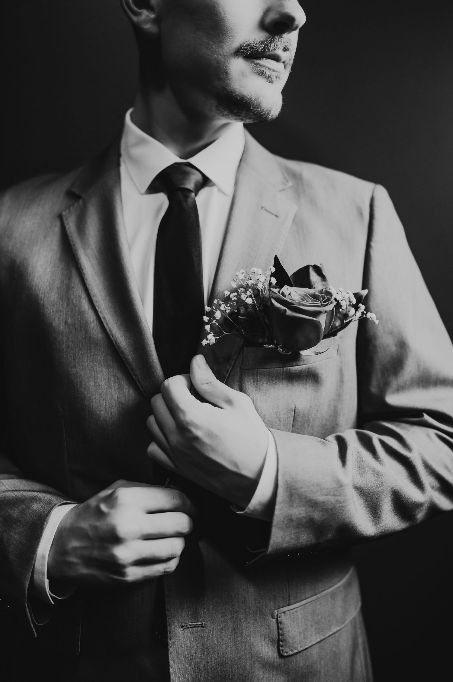 Black and white photo of a groom in a suit adjusting his boutonniere with a rose and baby's breath.
