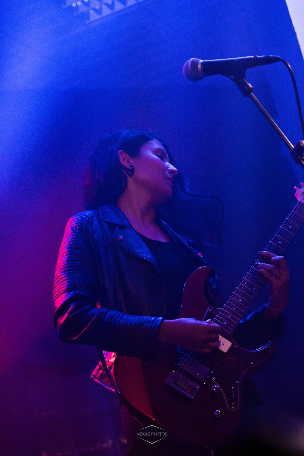 Female guitarist performing on stage under blue and red lights, playing electric guitar near a microphone.