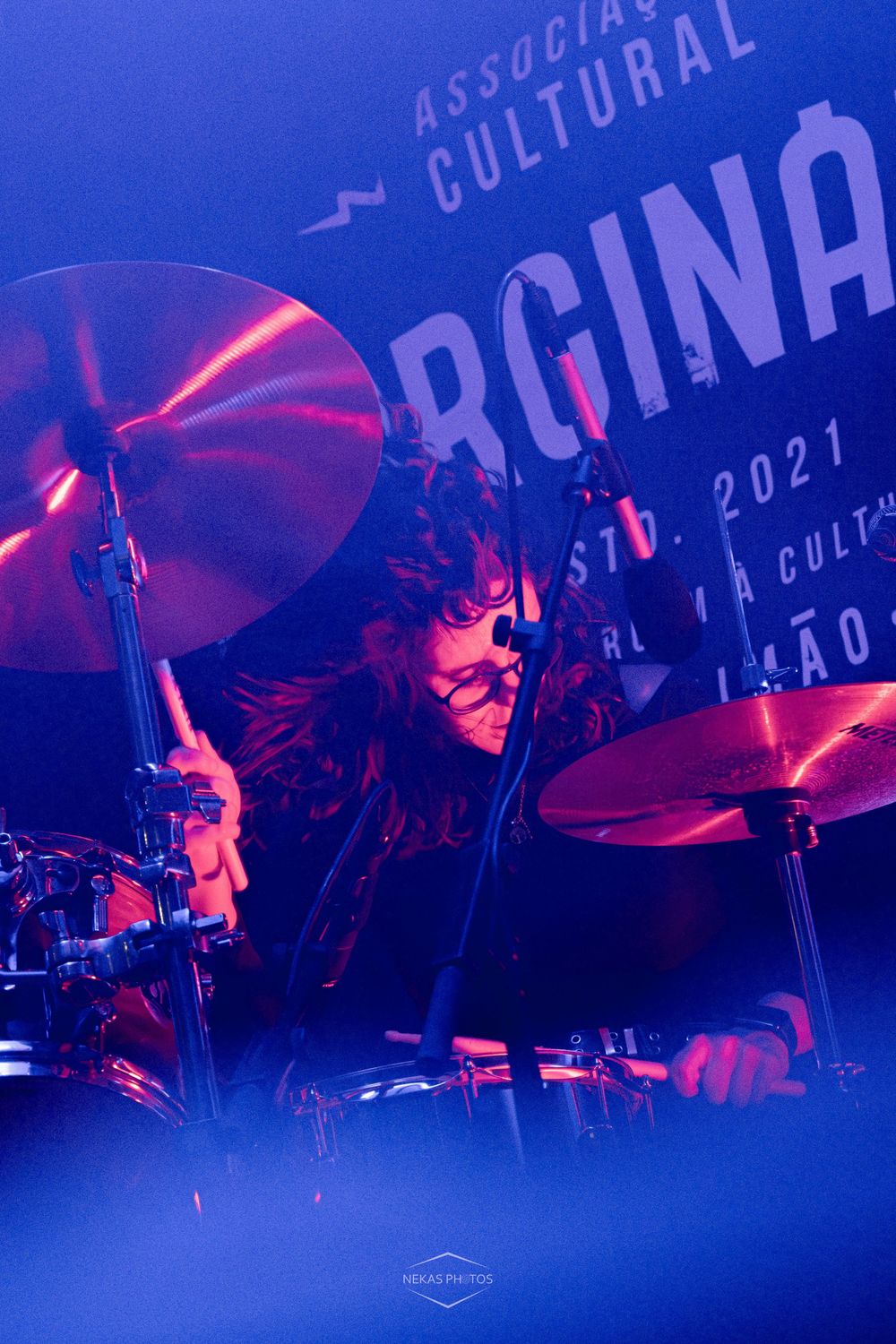 A passionate drummer performs energetically on stage bathed in blue and red lights at Associação Cultural in 2021.