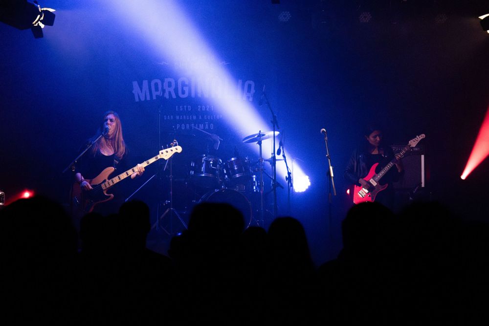 Band performing on stage with blue lighting, two guitarists and drummer visible at a live music venue.