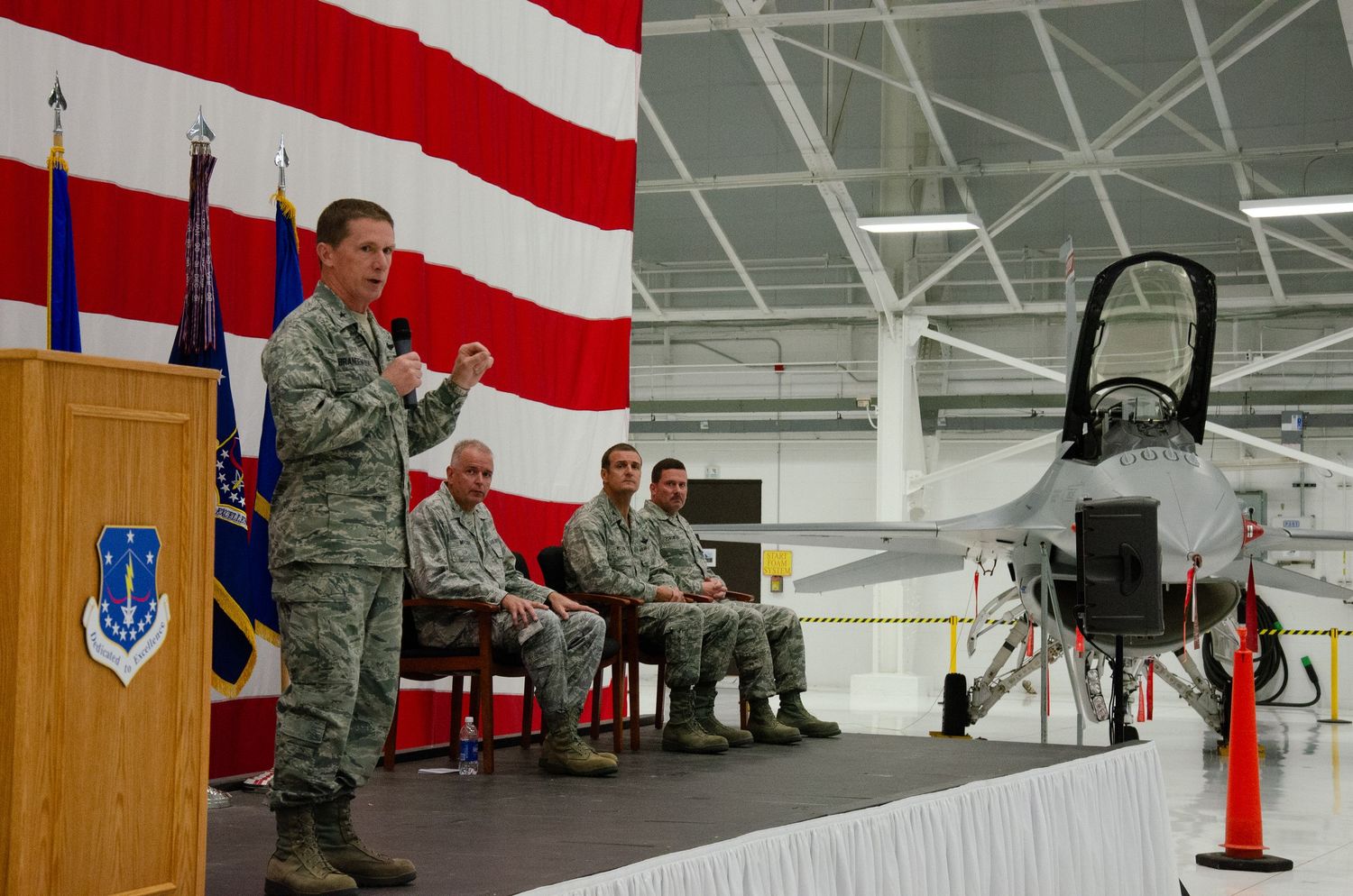 Military officer speaks at podium in hangar with F-16 jet, US flag backdrop, and seated uniformed personnel during ceremony.