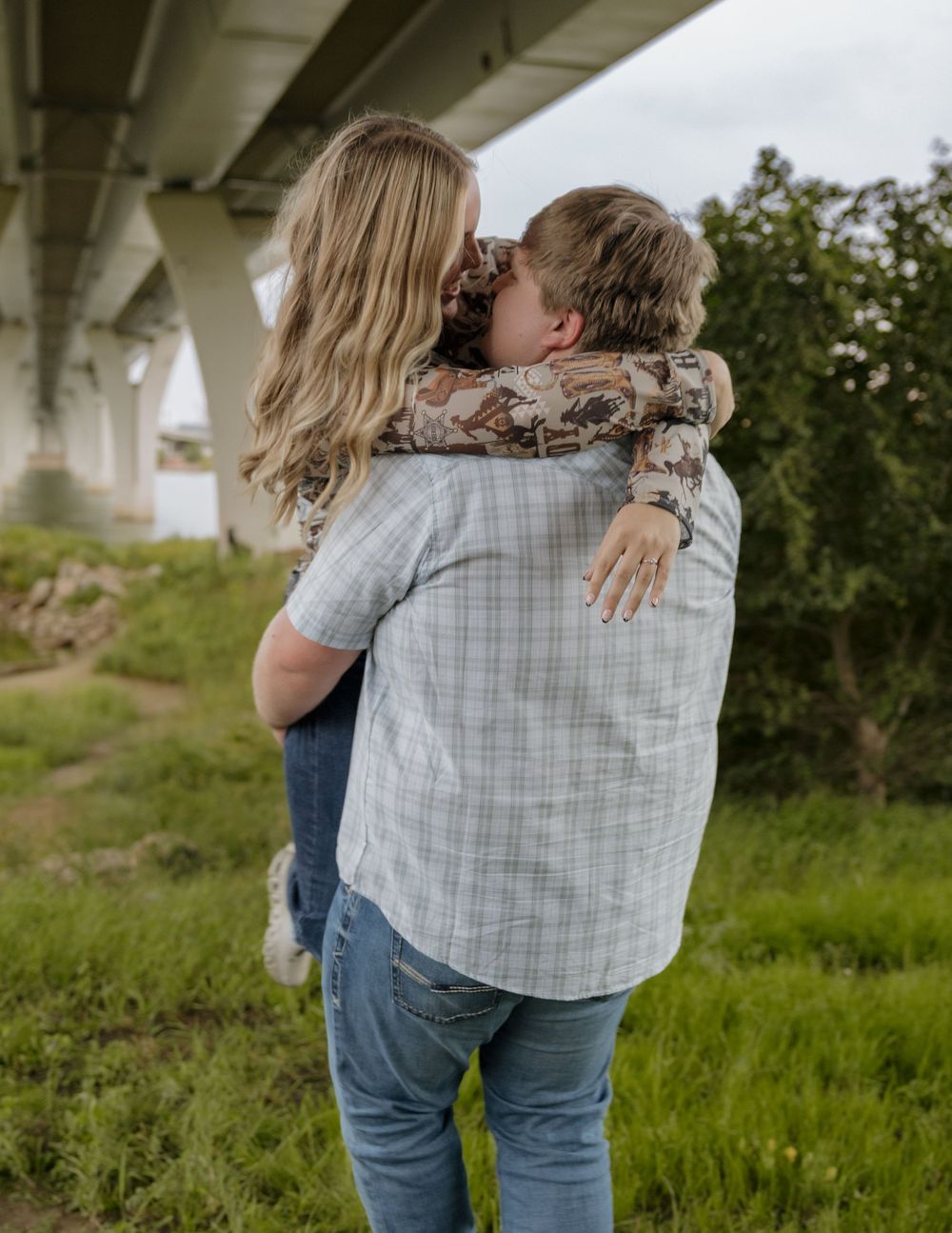 A couple shares a romantic kiss under a bridge, with the man lifting the woman in a loving embrace outdoors.