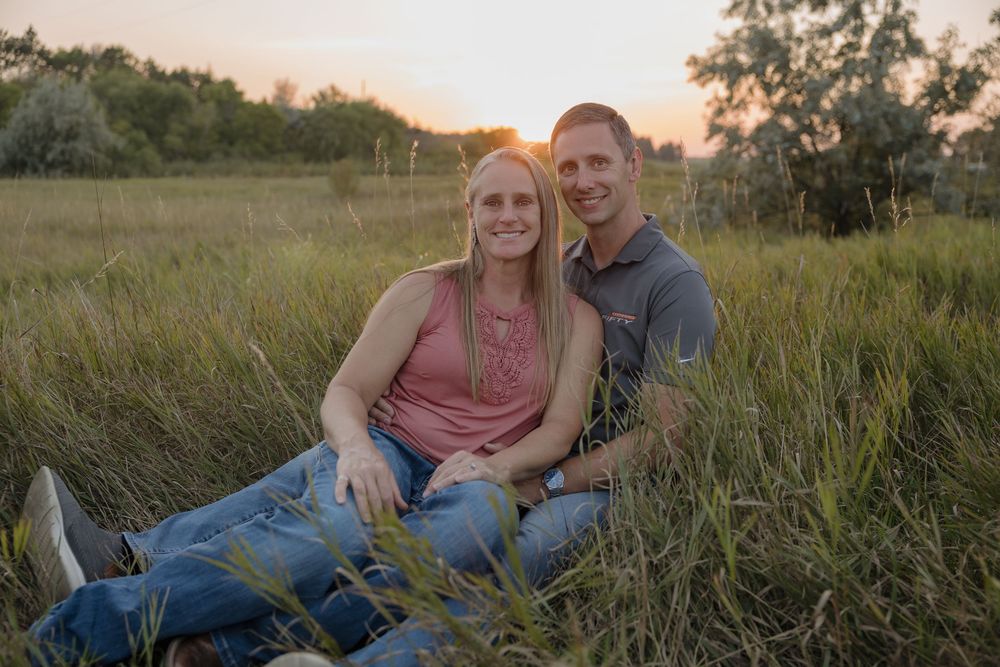 Couple sitting together in tall green grass field at golden sunset, smiling and embracing in a romantic outdoor portrait.