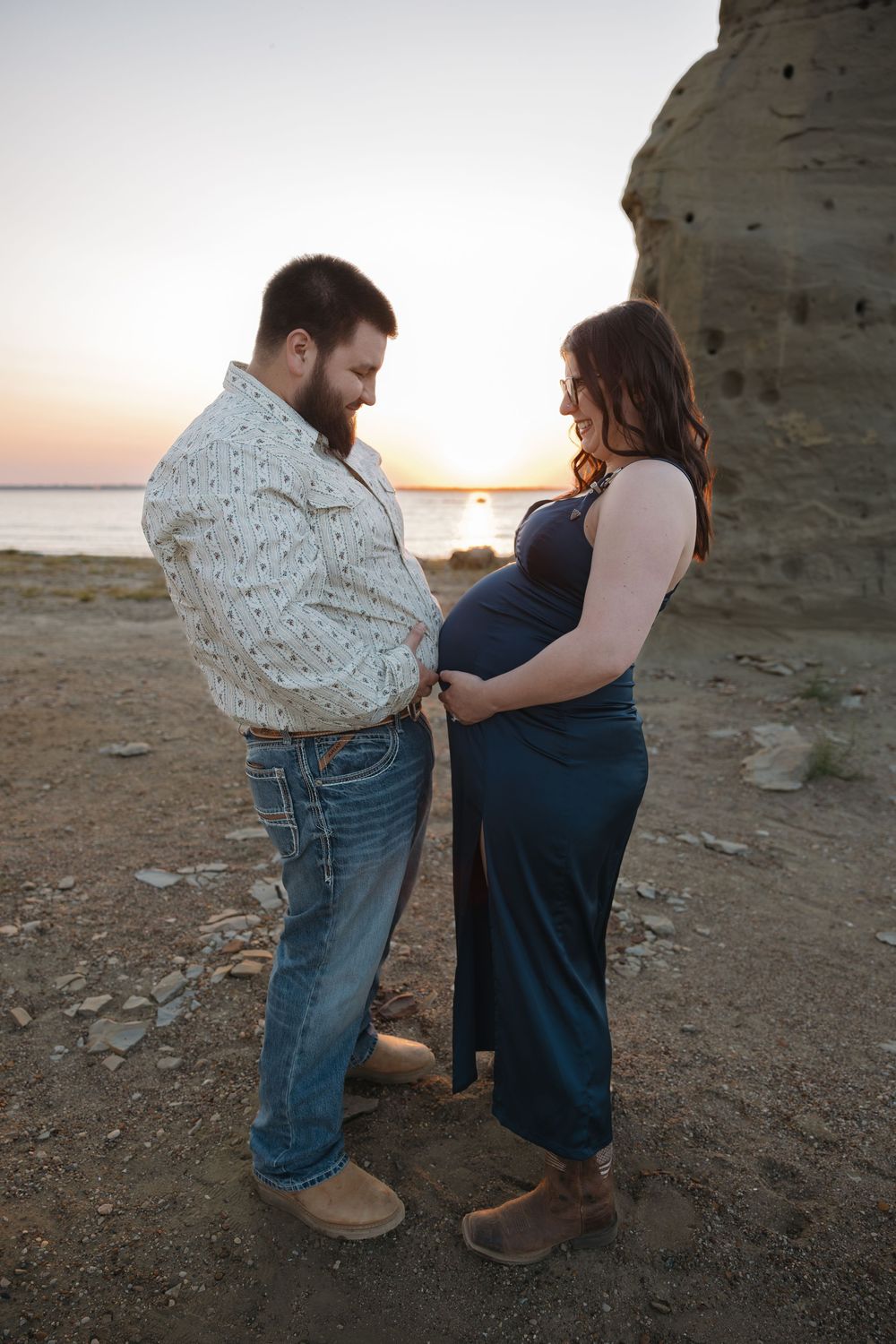 Couple sharing a tender moment at sunset on a rocky beach, man looking at pregnant woman's belly in blue maternity dress.
