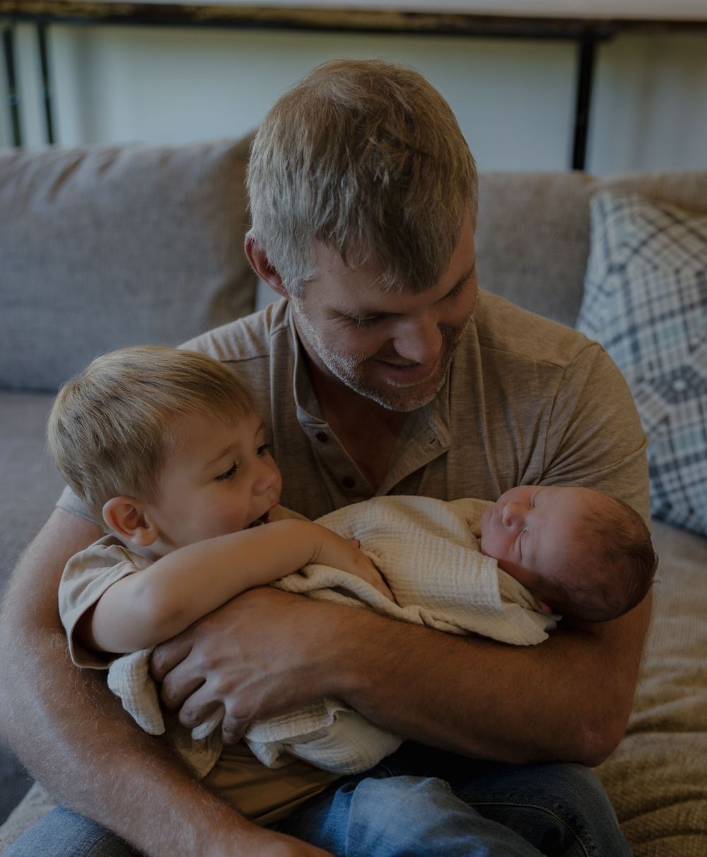 A father sits on a couch smiling at his newborn baby while a young boy lovingly kisses the infant.
