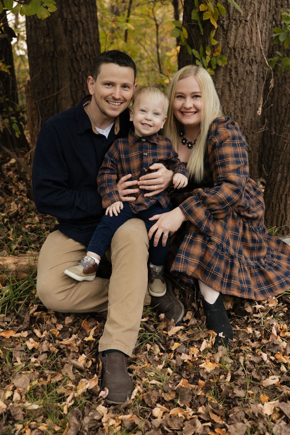 Family of three posing among autumn leaves in coordinating plaid outfits for a fall portrait session in the woods. Bismarck, ND photographer