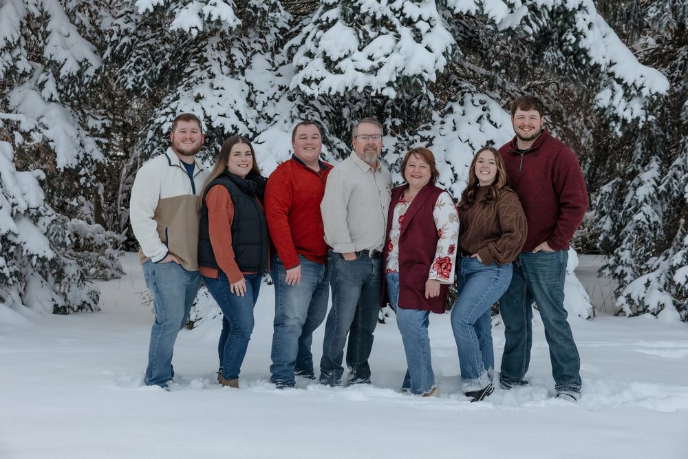 Seven people smiling together in a snowy winter landscape with snow-covered evergreen trees in the background. Bismarck, ND photographer
