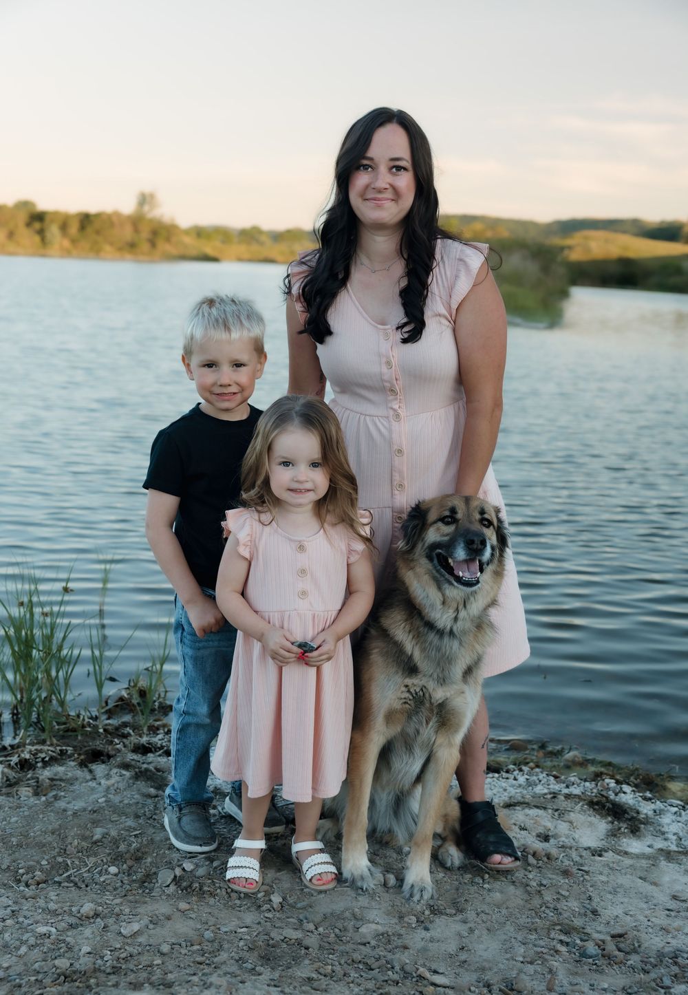 A smiling woman stands with two young children and a large dog near a scenic lakeside at sunset. Bismarck, ND photographer