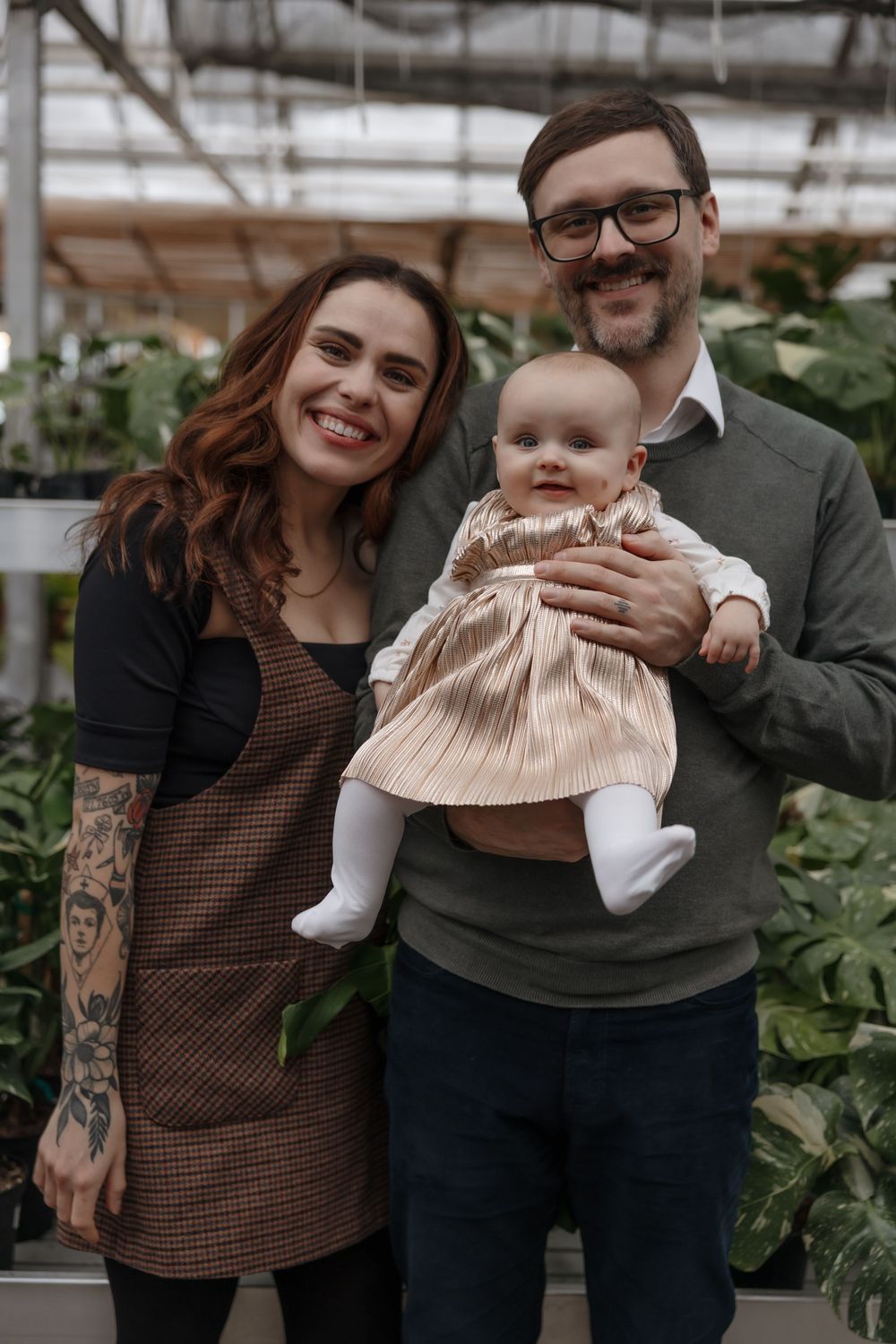 Smiling family with tattooed mother, bearded father wearing glasses, and baby girl in pink dress posing in a greenhouse. Bismarck, ND photo
