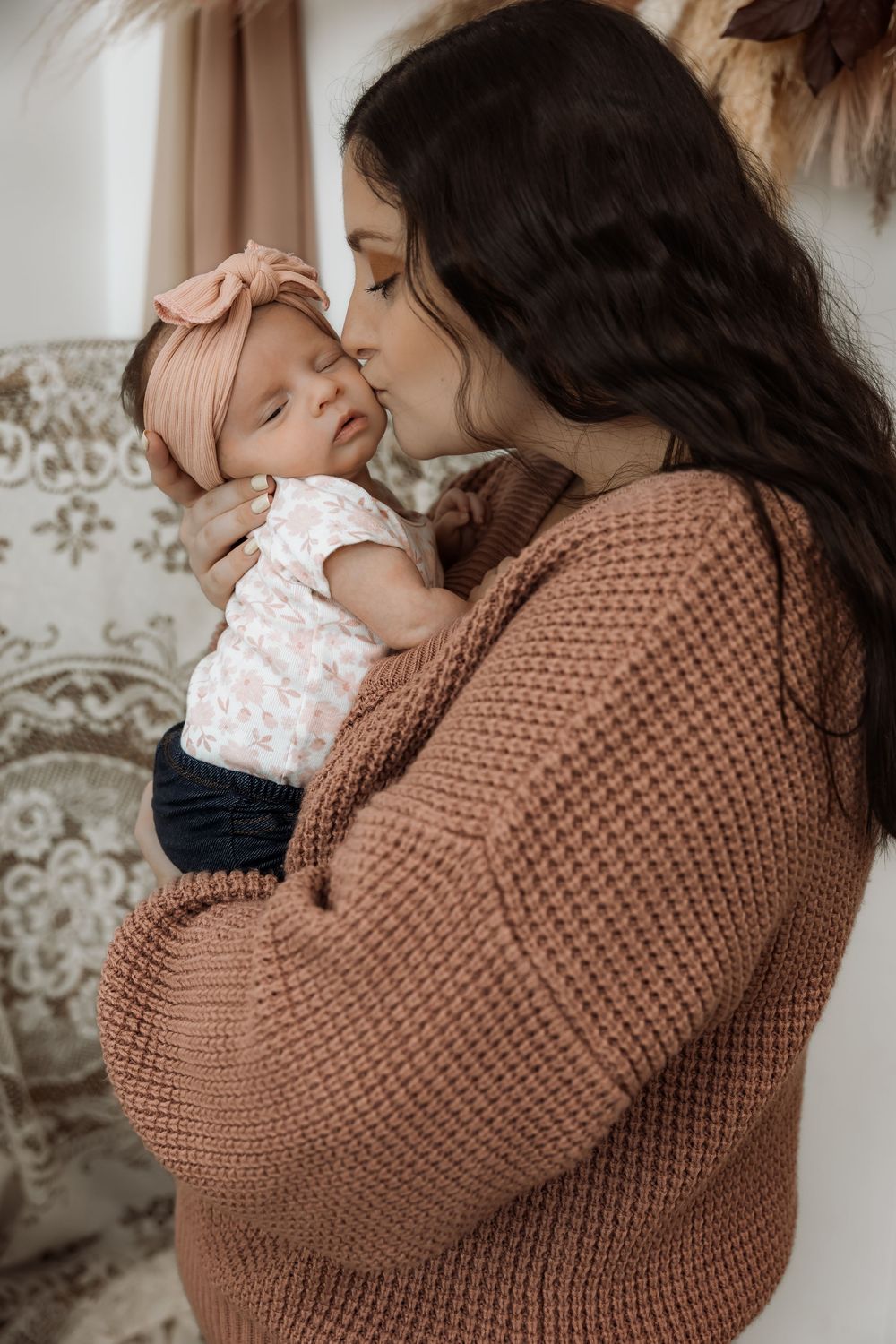 A mother in a brown knit sweater tenderly kisses her sleeping newborn baby girl wearing a pink bow headband. Bismarck, ND photographer