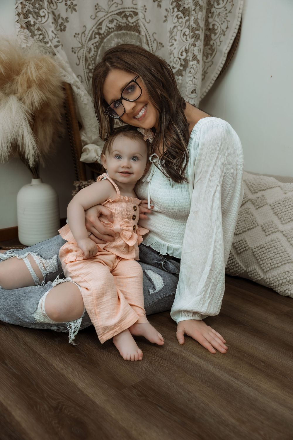 Young woman with glasses sits on floor holding a baby girl in peach romper in a boho-styled room. Bismarck, ND photographer