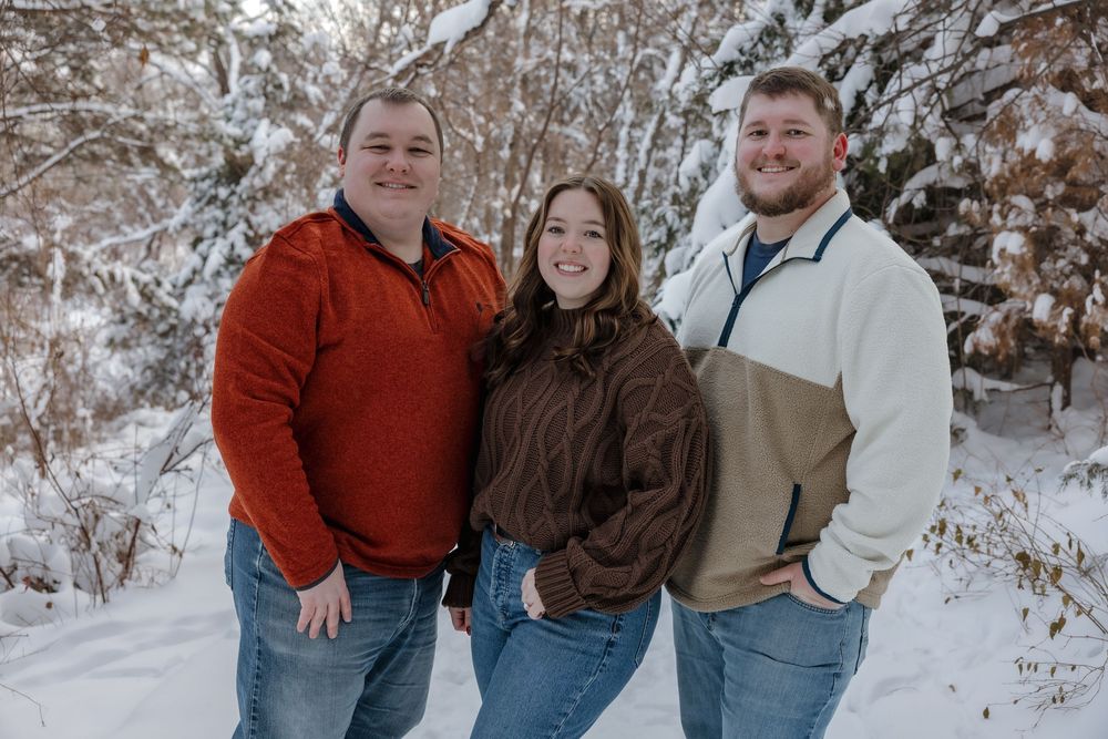 Three adults smiling together in a snowy winter forest, wearing colorful sweaters and jeans. Bismarck, ND photographer