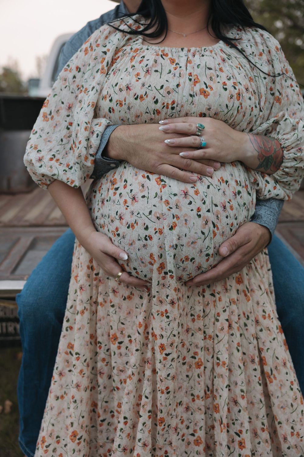 Pregnant woman in floral dress cradling baby bump, partner embracing her from behind outdoors. Garrison, ND Photographer