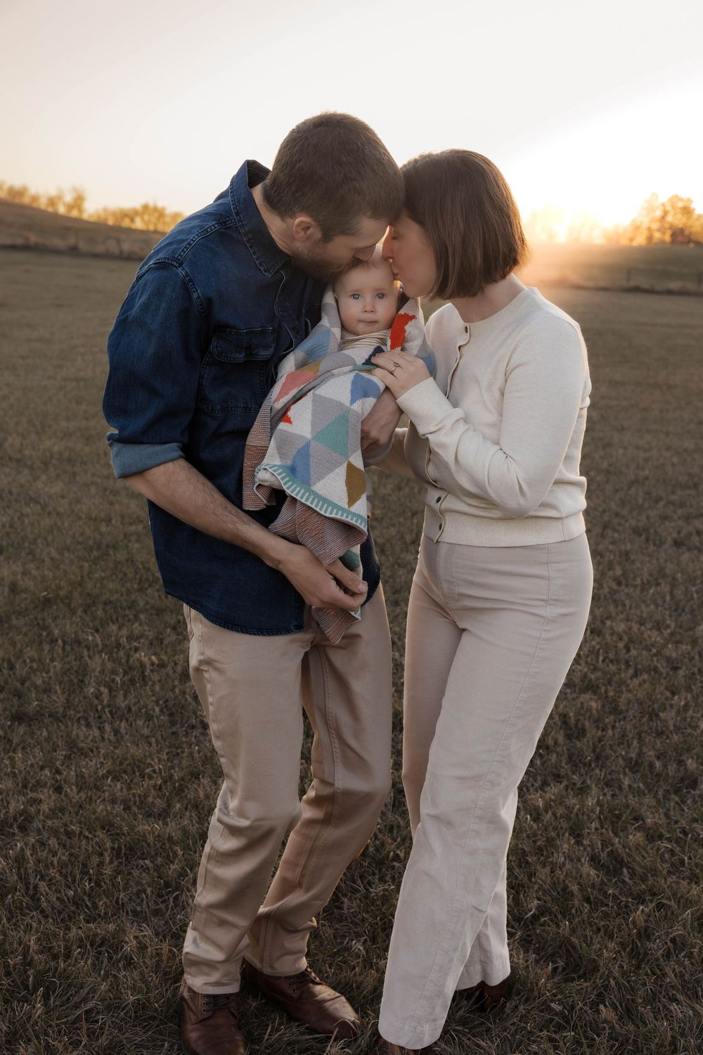 A young family stands in a golden sunset field, parents lovingly kissing their baby wrapped in a colorful blanket. Garrison, ND Photographer