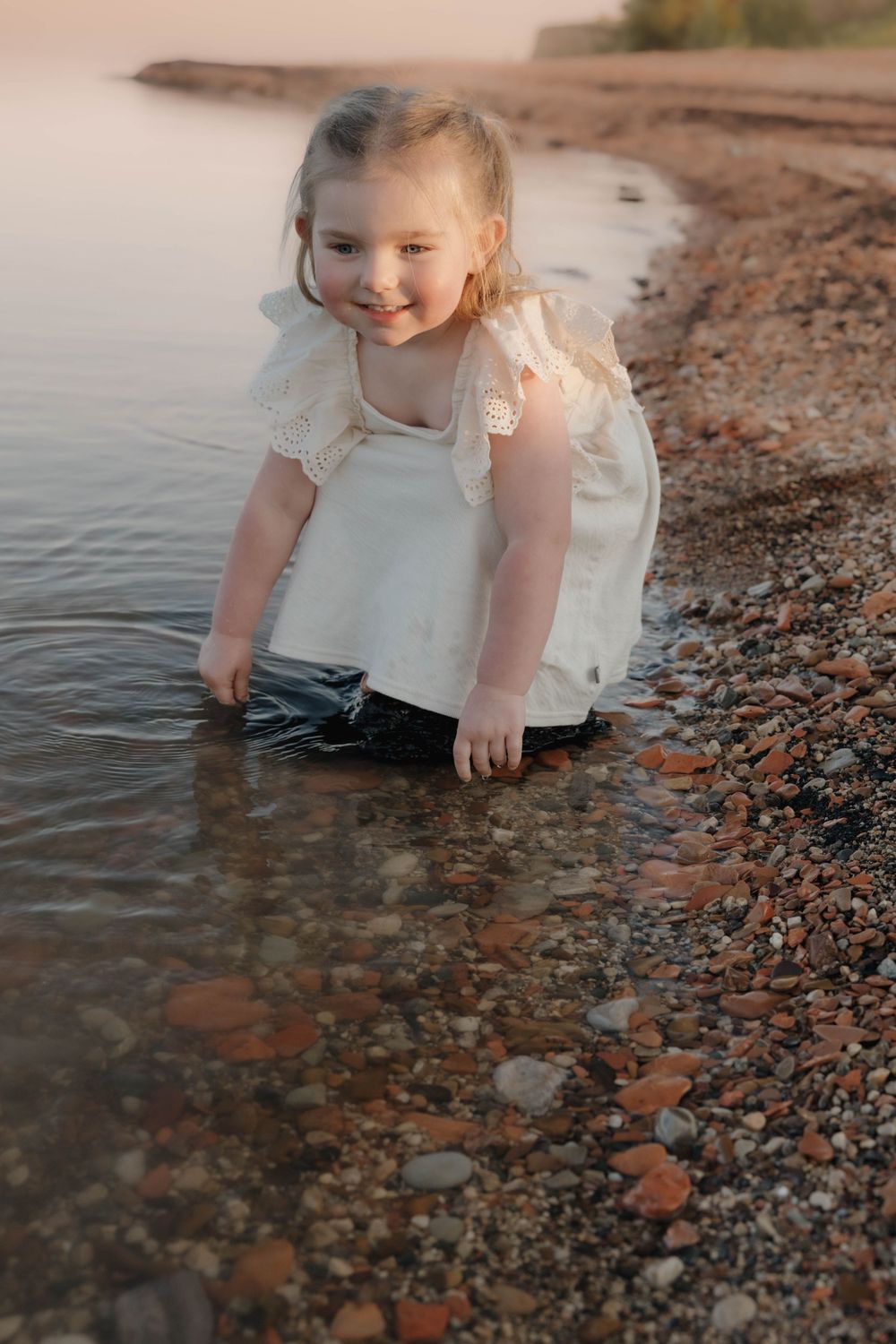 A smiling little girl in a white dress playing in shallow water on a rocky shoreline at sunset. Garrison, ND Photographer