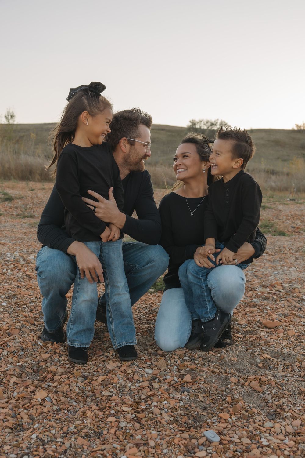 Family of four in matching black tops and jeans laughing together outdoors on a rocky landscape. Garrison, ND Photographer