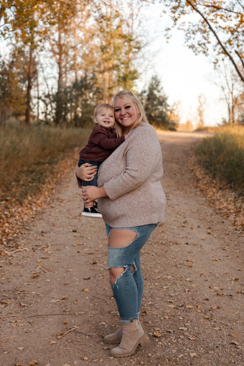 Pregnant woman holding toddler on autumn dirt path, wearing ripped jeans and tan sweater, smiling outdoors. Garrison, ND Photographer