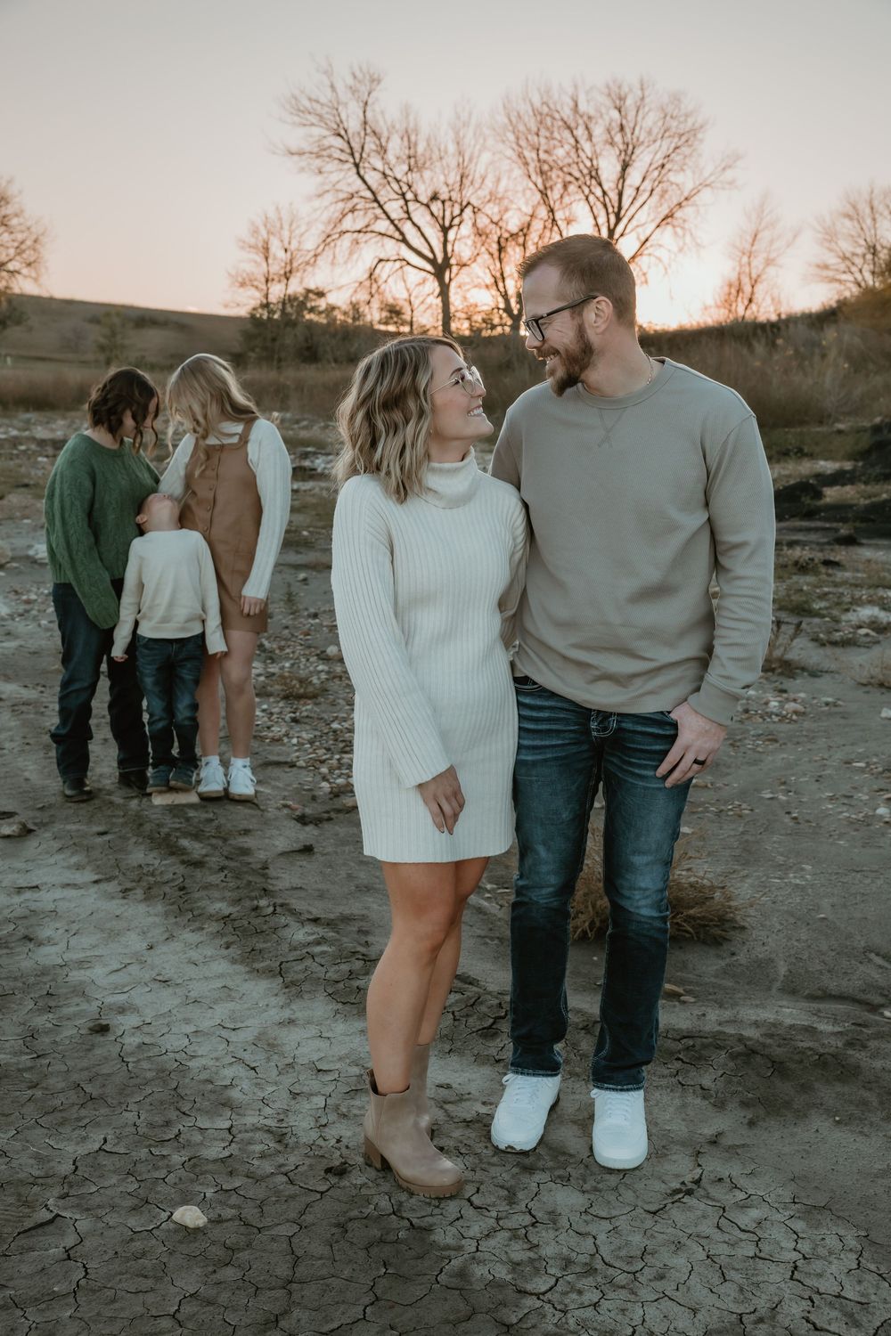 Couple smiling at each other outdoors at sunset with children playing behind them. Garrison, ND Photographer