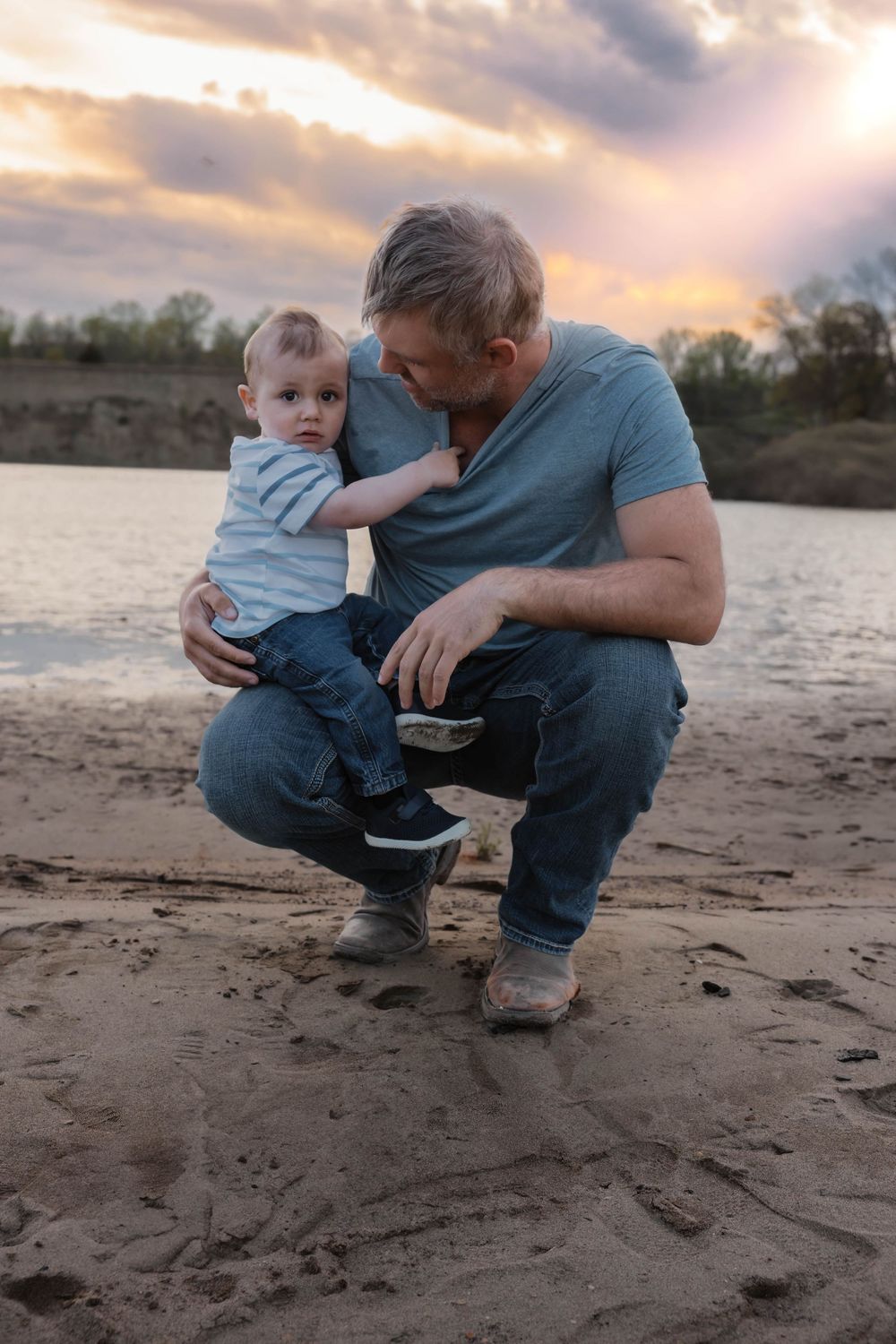 Father crouching on sandy beach holding toddler at sunset with river in background. Garrison, ND Photographer