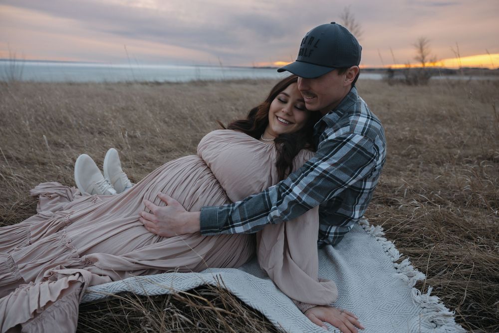 Couple sharing a tender maternity moment in a field at sunset, woman in pink dress, man in plaid shirt and cap. Garrison, ND Photographer