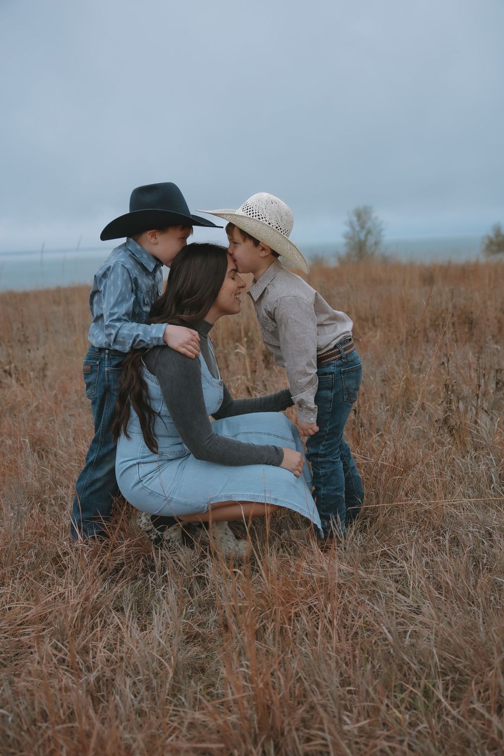 A mother kneels in a golden field as two young boys in cowboy hats lean in to kiss her cheeks. Garrison, ND Photographer