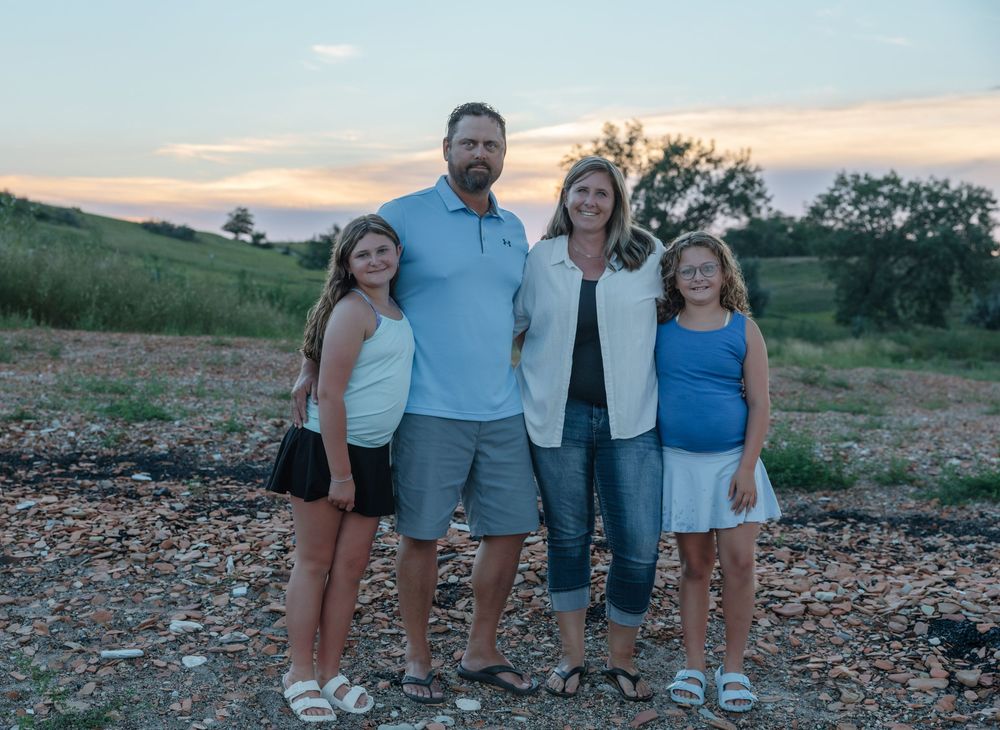 Family of four poses outdoors at sunset on rocky ground with green hills and colorful sky in background. Garrison, ND Photographer
