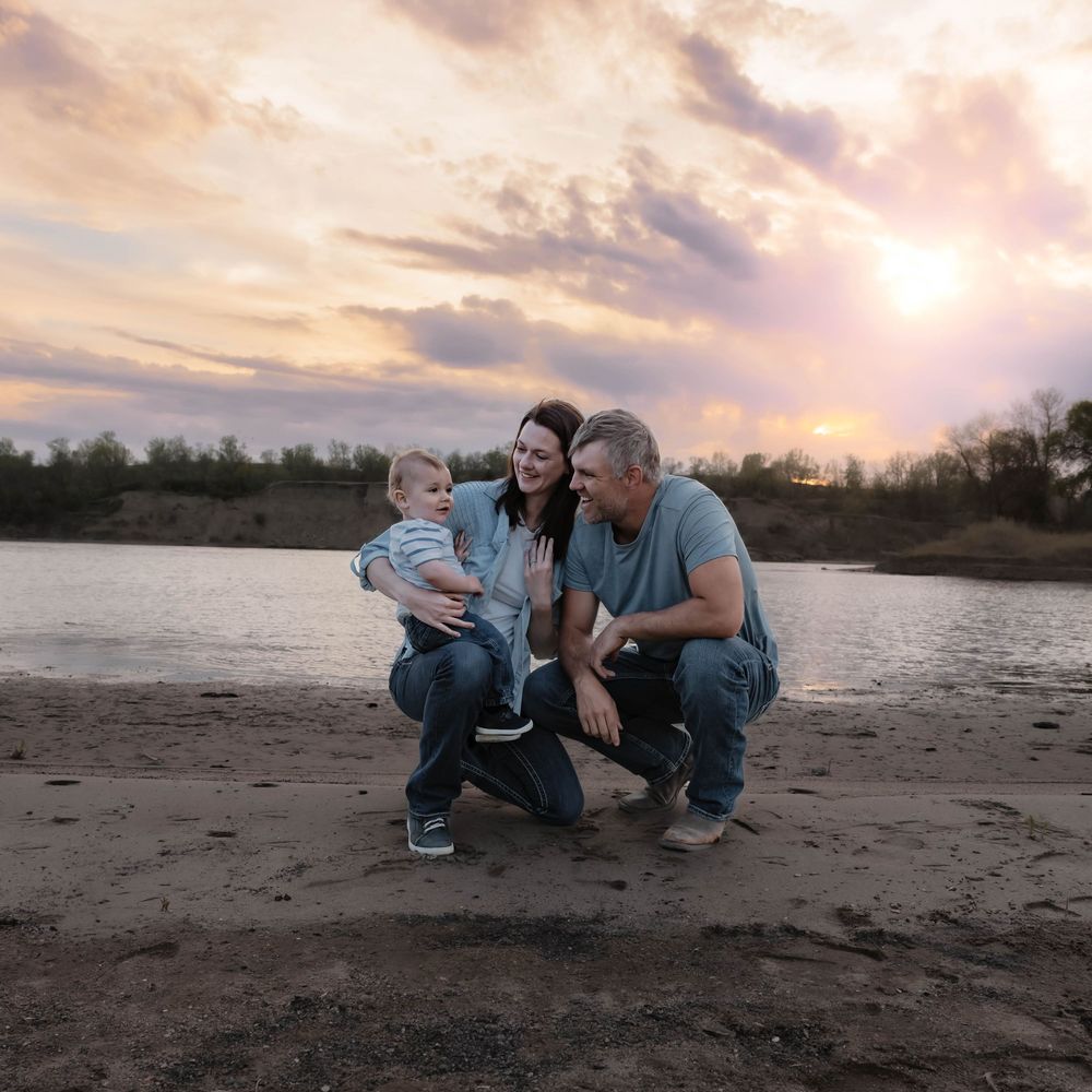 Family of three posing by a river at sunset, parents smiling at baby on sandy shore under dramatic cloudy sky. Garrison, ND Photographer