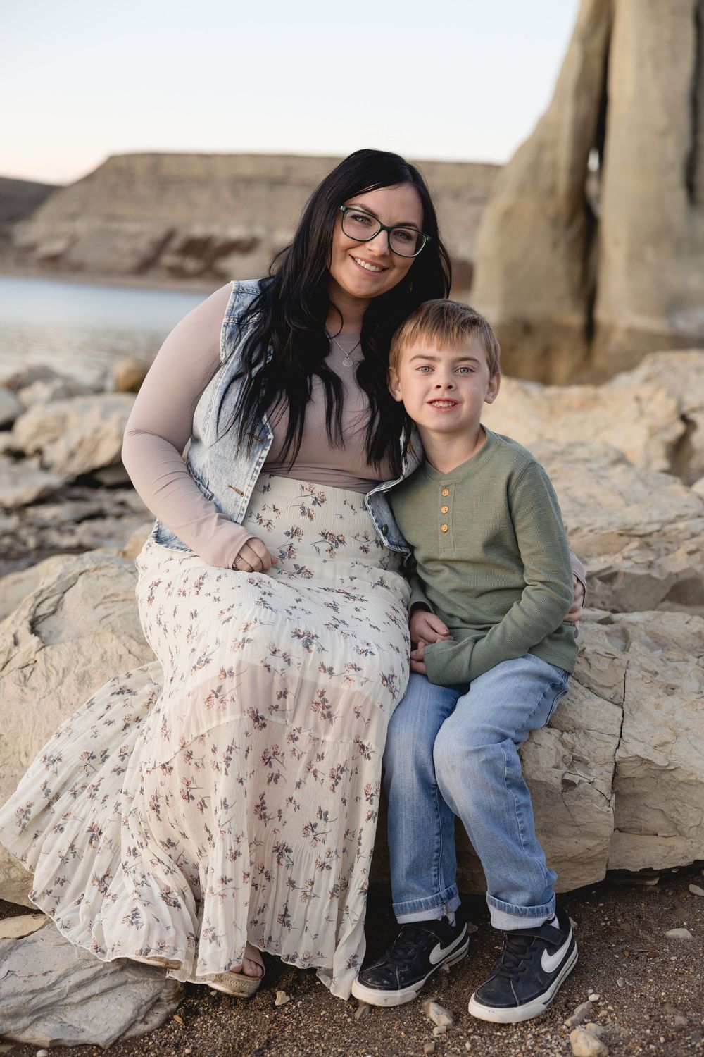 A mother with dark hair and glasses sits on rocks with her young son in a green shirt, smiling outdoors. Garrison, ND Photographer