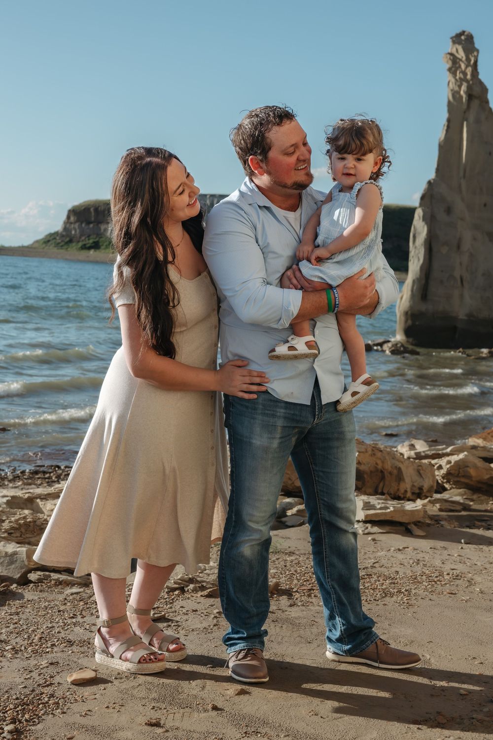 Family of three posing on rocky beach, man holding toddler with woman beside them, ocean and cliffs in background. Garrison, ND Photographer
